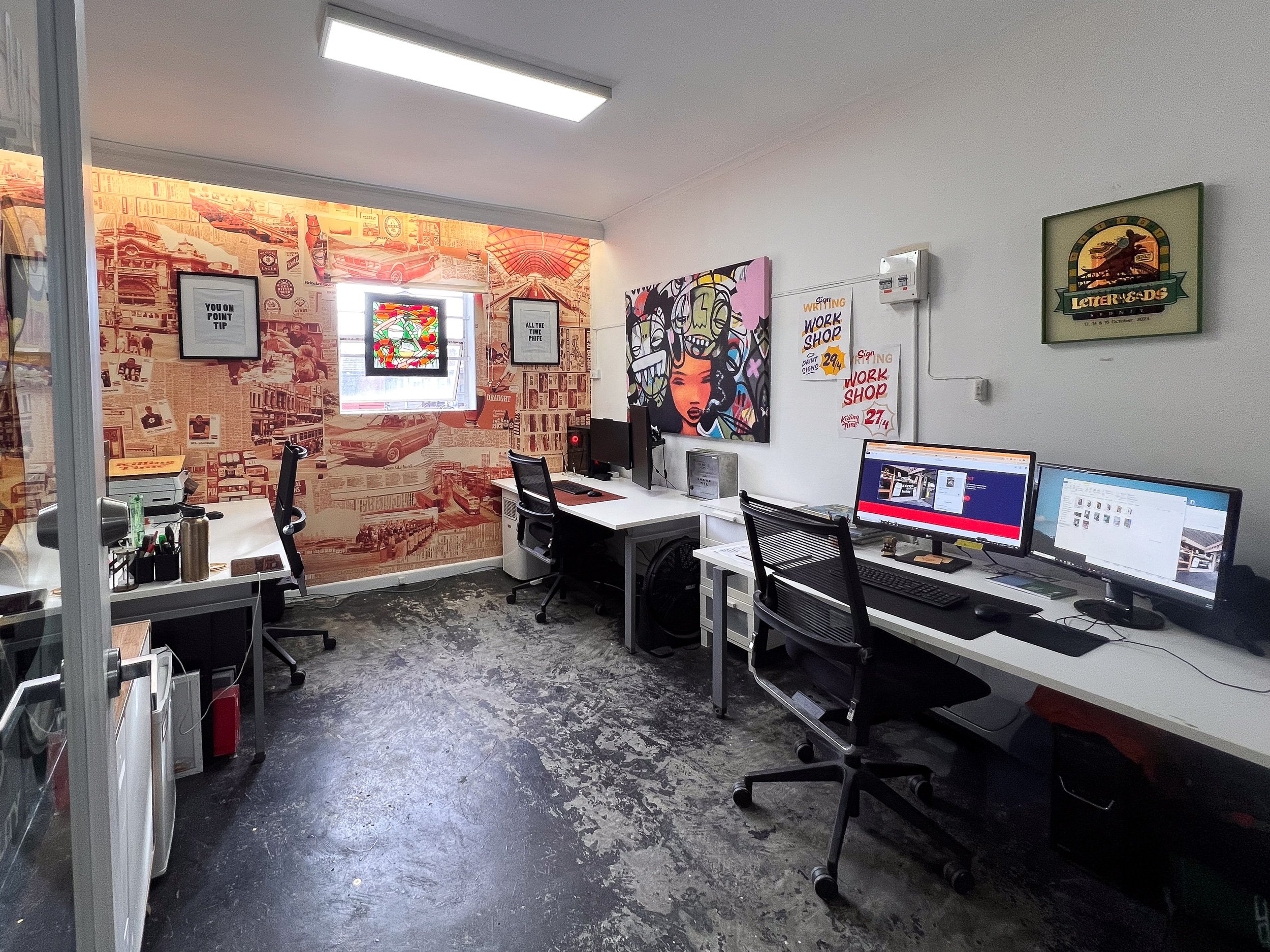 An office workspace with two desks, three computer monitors, two chairs, and wall decorations including statement posters and artwork. The floor is worn, and there is a small window with colorful stained glass.