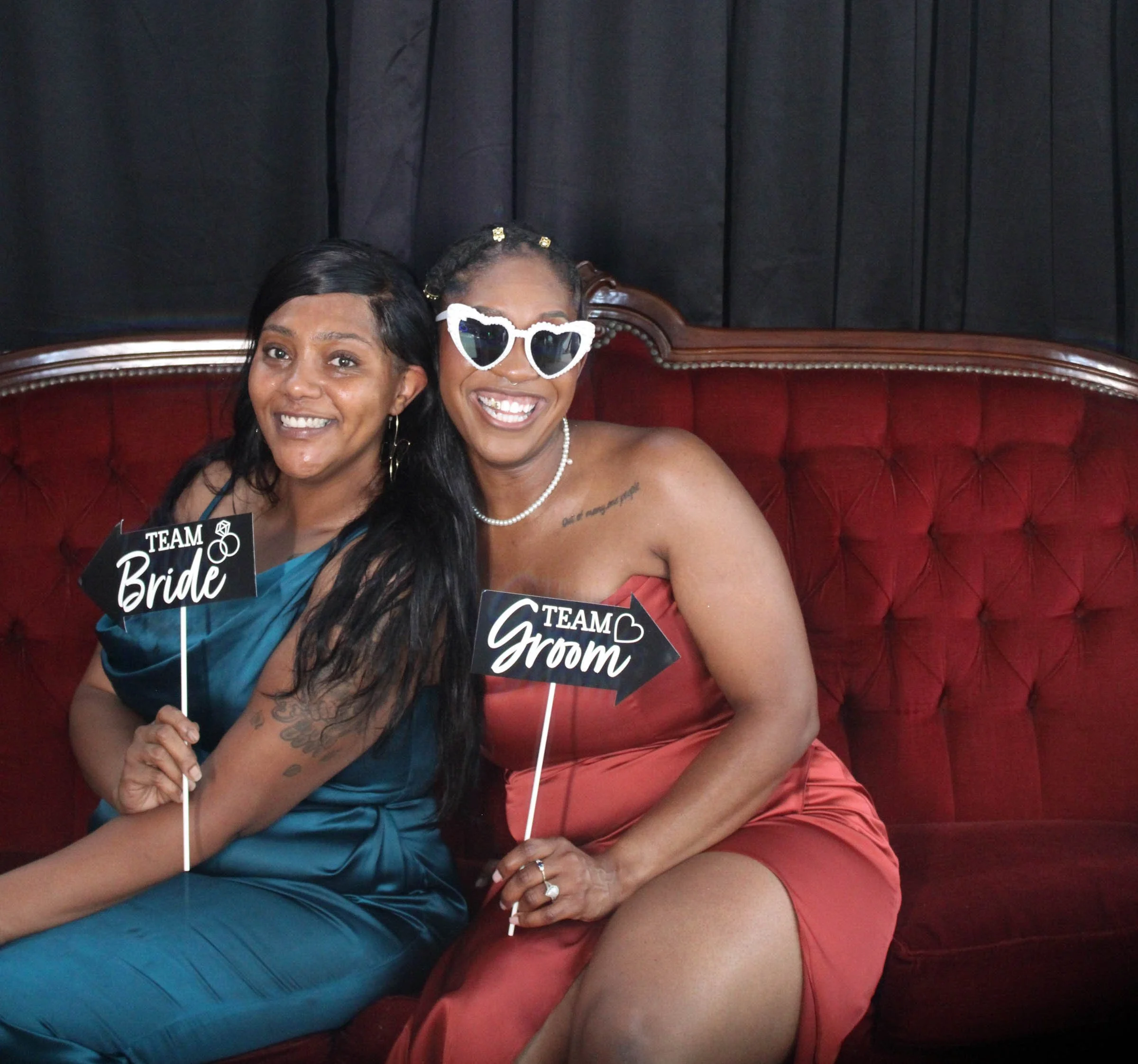 Two women sitting on a red vintage couch, smiling and holding signs that read 'Team Bride' and 'Team Groom', with one woman wearing heart-shaped sunglasses.