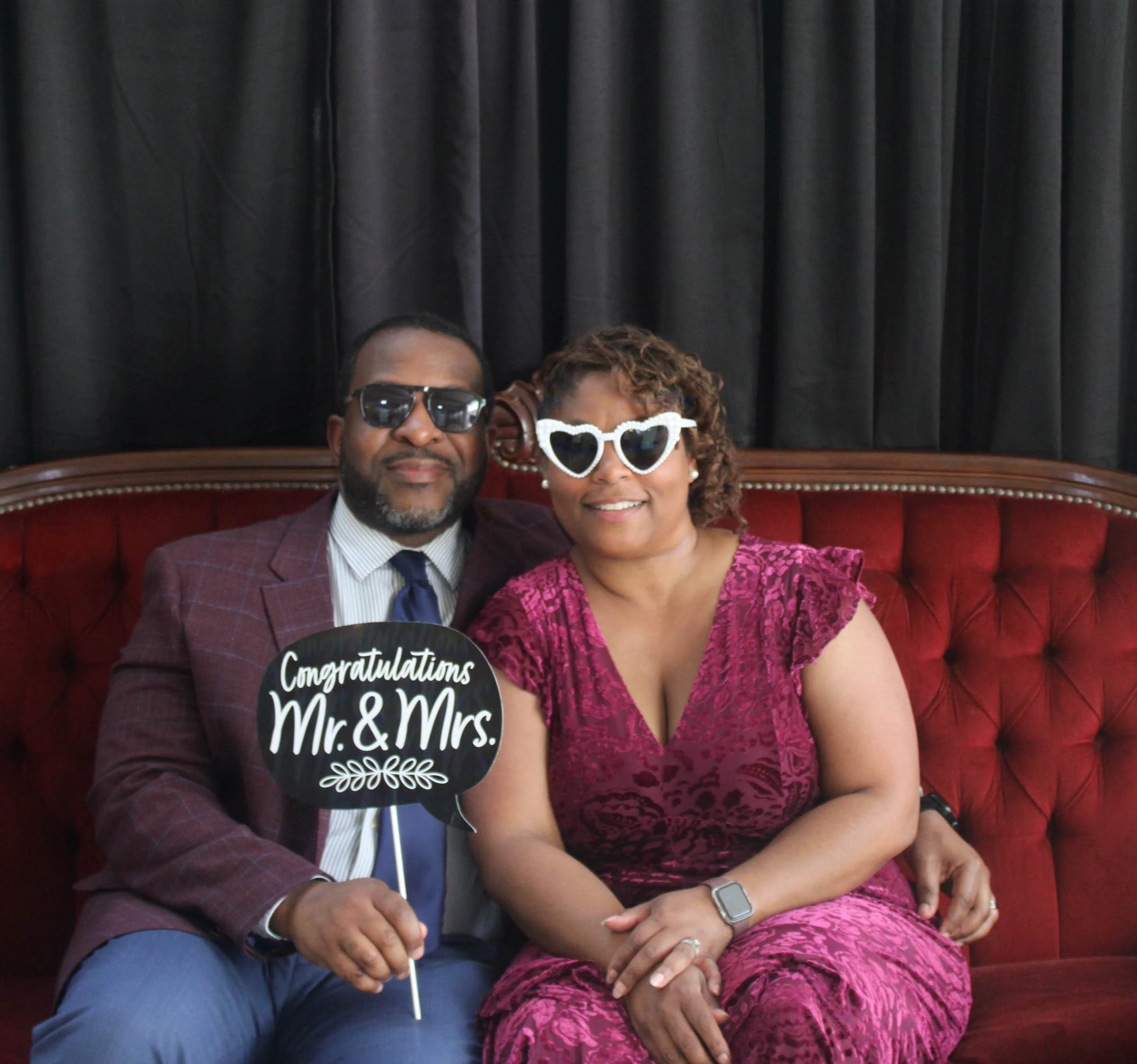 A group of people sitting on a red velvet bench with black curtains behind. A man wearing sunglasses and a woman wearing silly heart-shaped glasses are smiling at the camera. The woman is holding a small black sign that says 'Congratulations Mr. & Mr