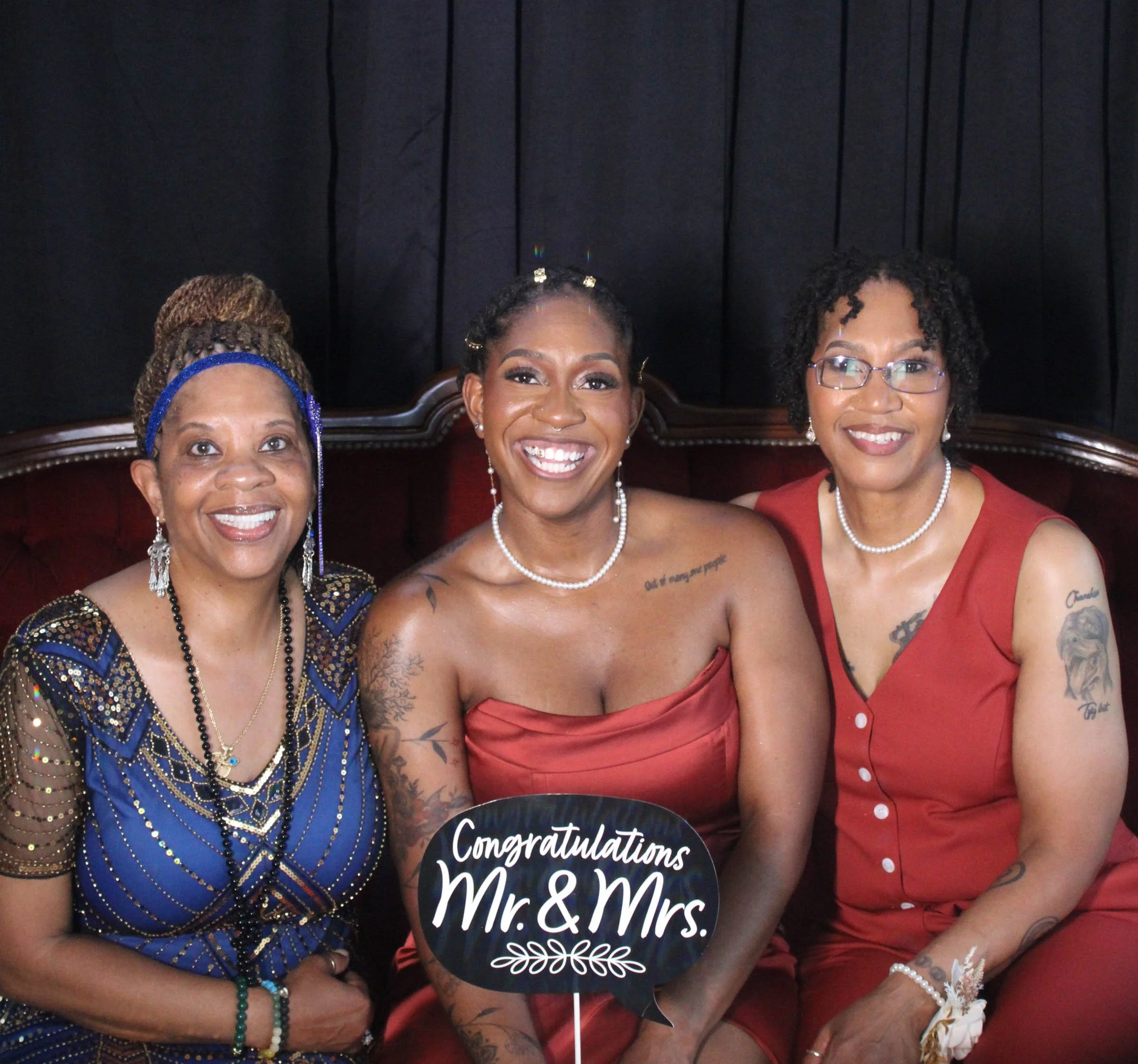 Celebration of a wedding, three women dressed in elegant attire holding a sign that reads 'Congratulations Mr. & Mrs.' in front of a dark backdrop.