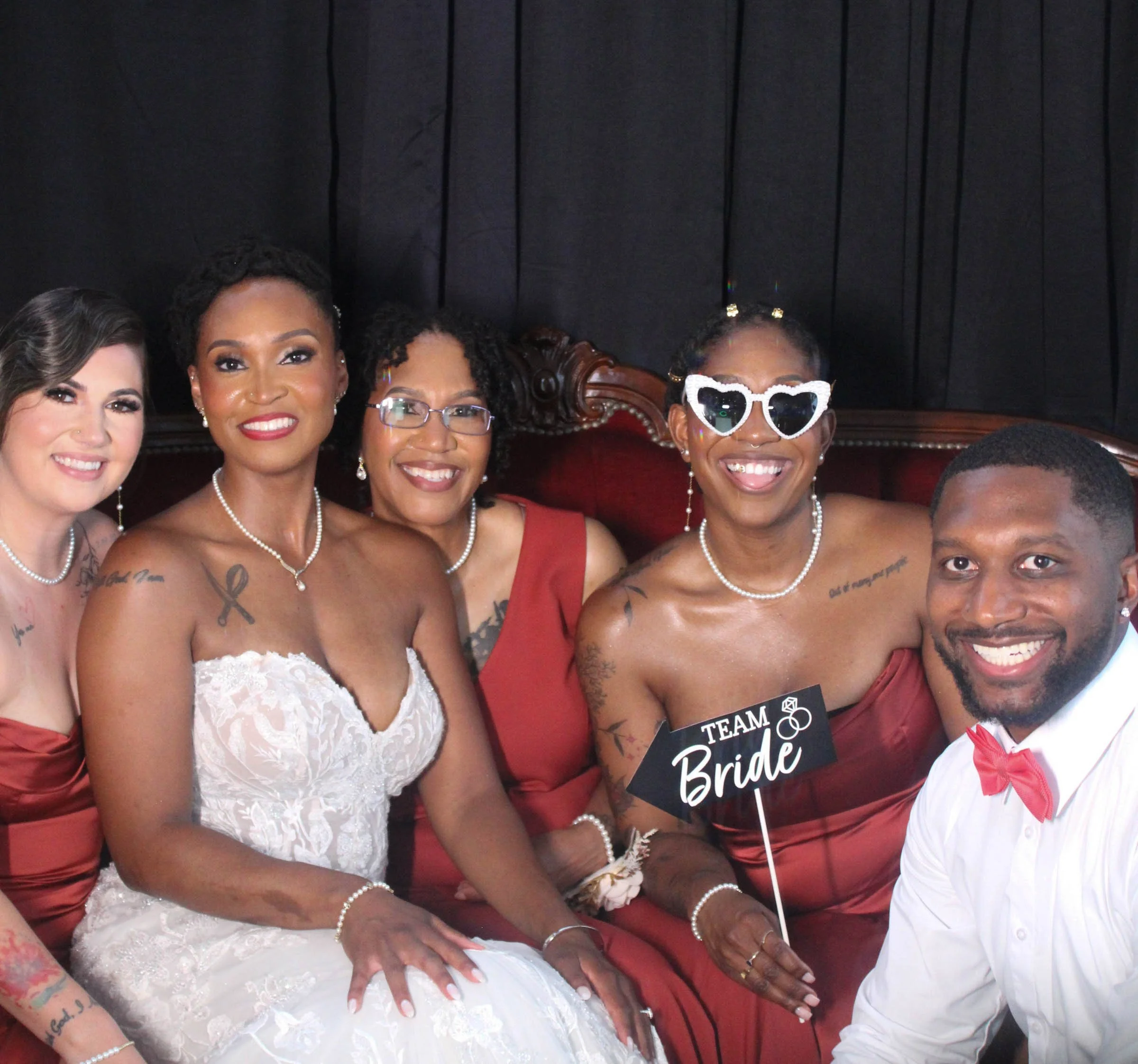 Group of smiling women dressed in elegant attire, celebrating at a wedding, with one woman holding a 'Team Bride' sign, standing against a black curtain background.