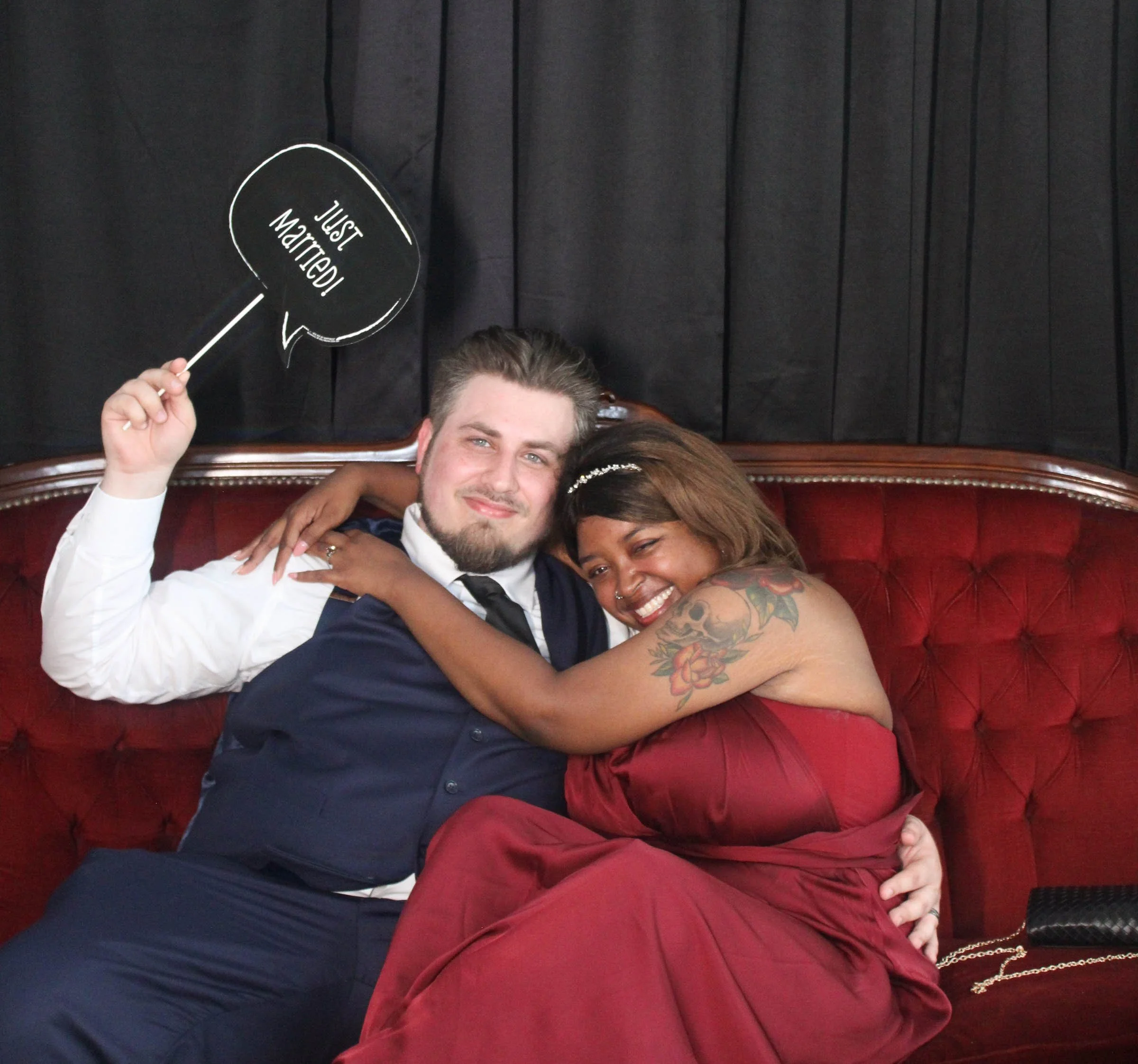 Close-up photo of happy couple smiling, hugging, with a man holding a speech bubble sign that says 'Just Married!', in an indoor setting with dark wall background.