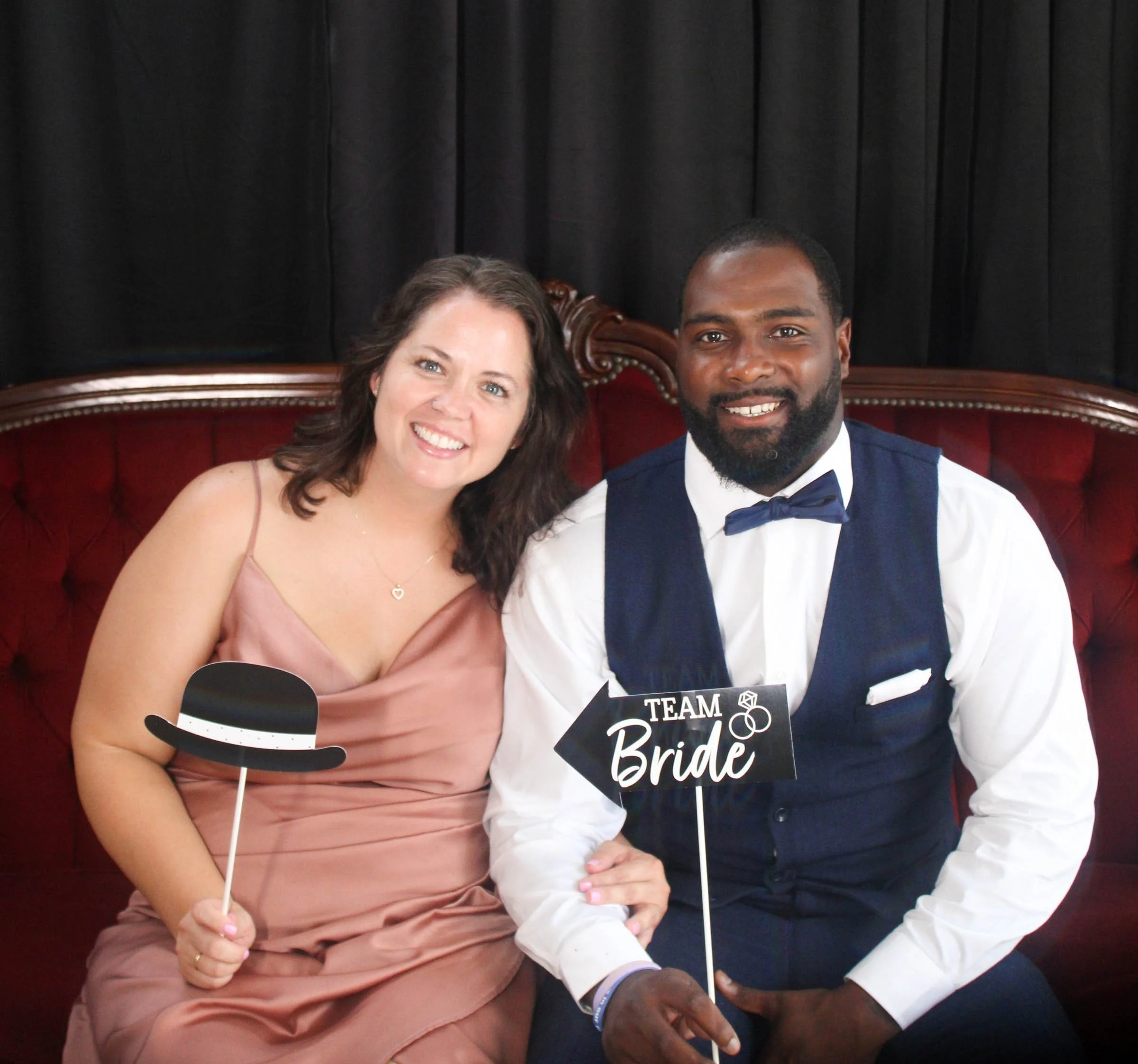 A bride and groom sitting on a red velvet couch, holding signs that say "TEAM Bride" and a mustache prop. They are smiling, with black curtains in the background.