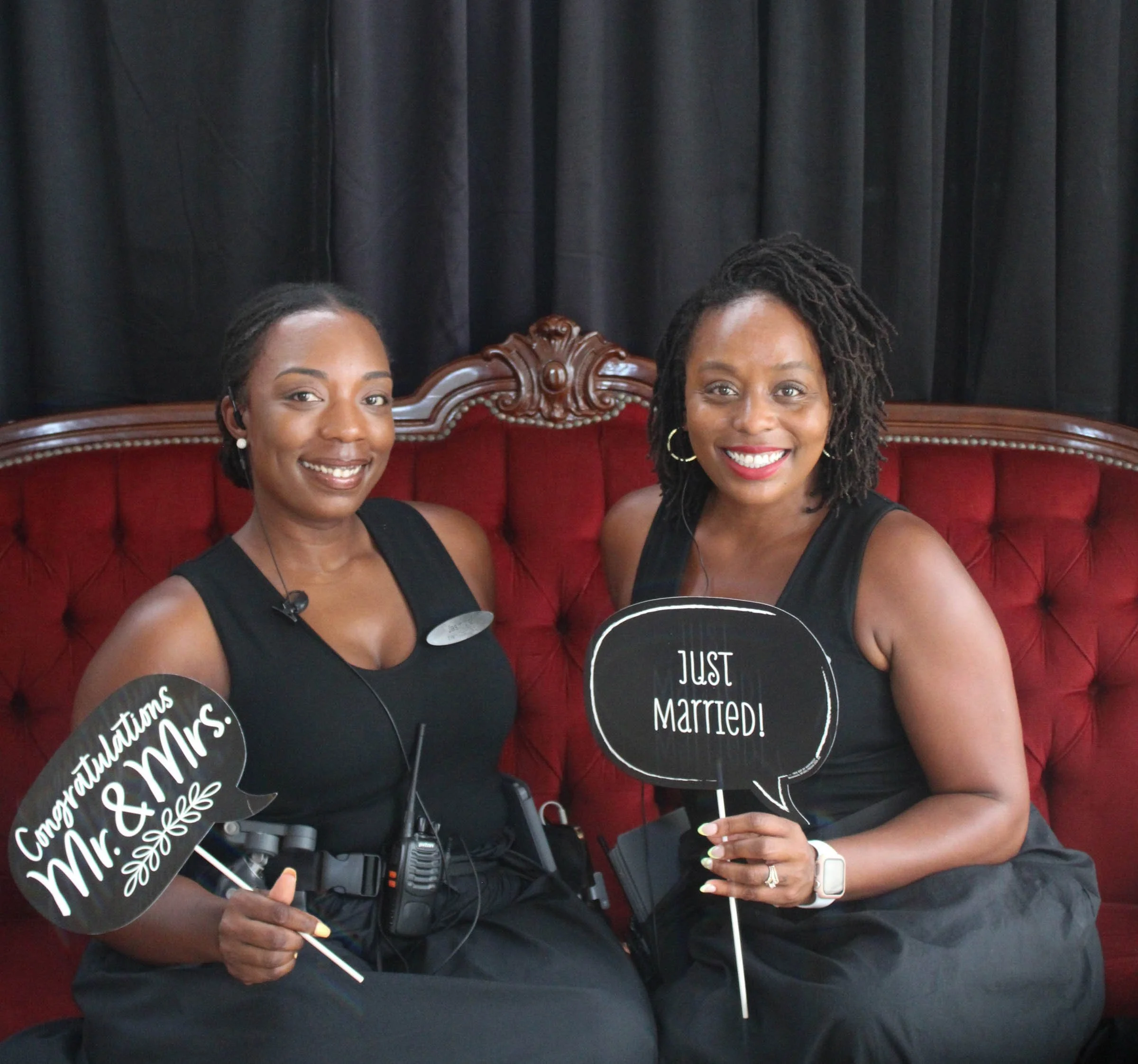 Two women sitting on a red velvet couch, smiling at the camera, holding speech bubble signs that read 'Congratulations Mr. & Mrs. Married!' and 'Just Married.' One woman wears a black dress and the other wears a black dress with a necklace. Both wome