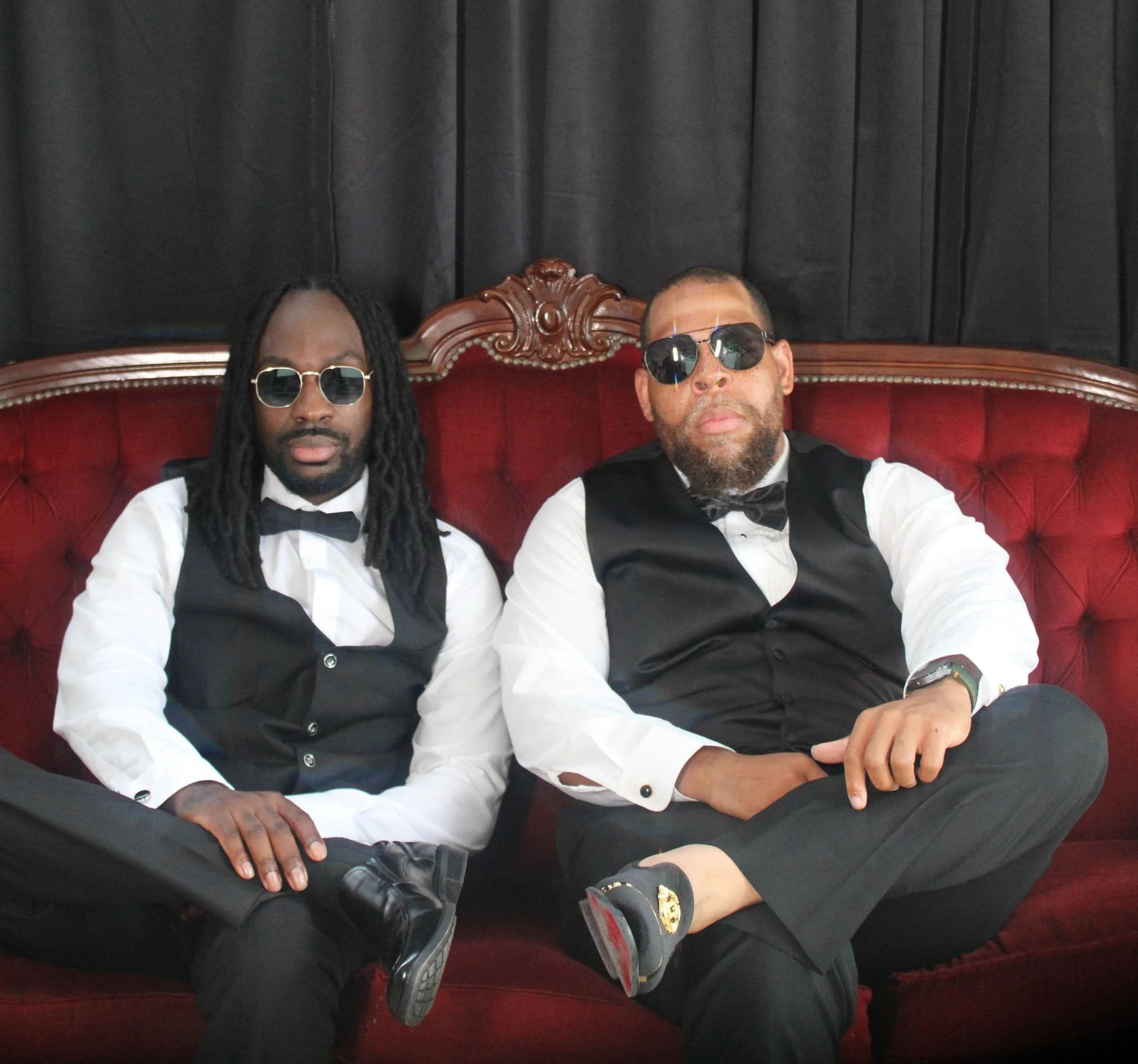 Two men dressed in tuxedos and sunglasses seated on a red velvet throne with black curtains in the background.