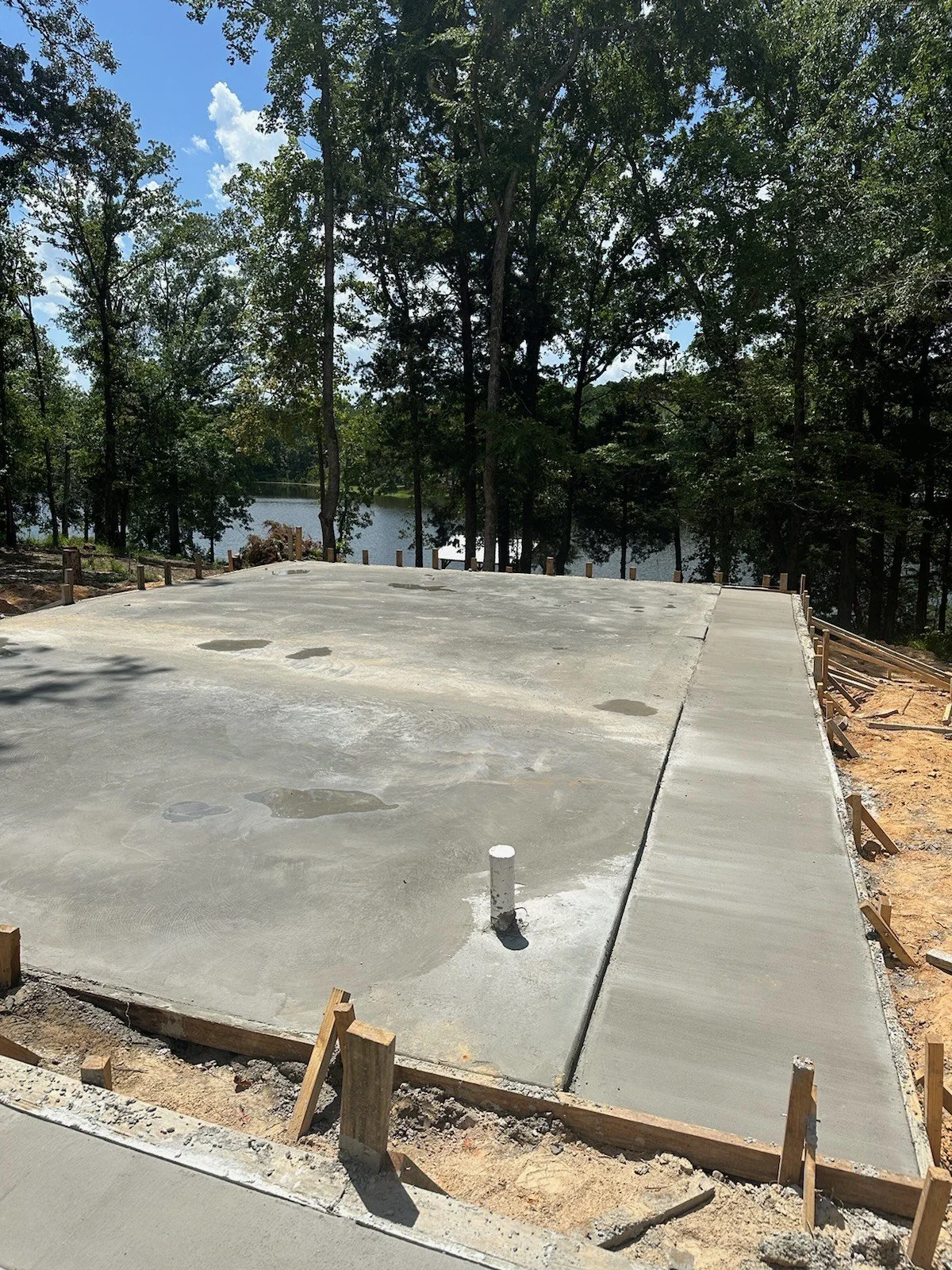 Freshly poured concrete slab on a construction site surrounded by trees with a lake in the background.