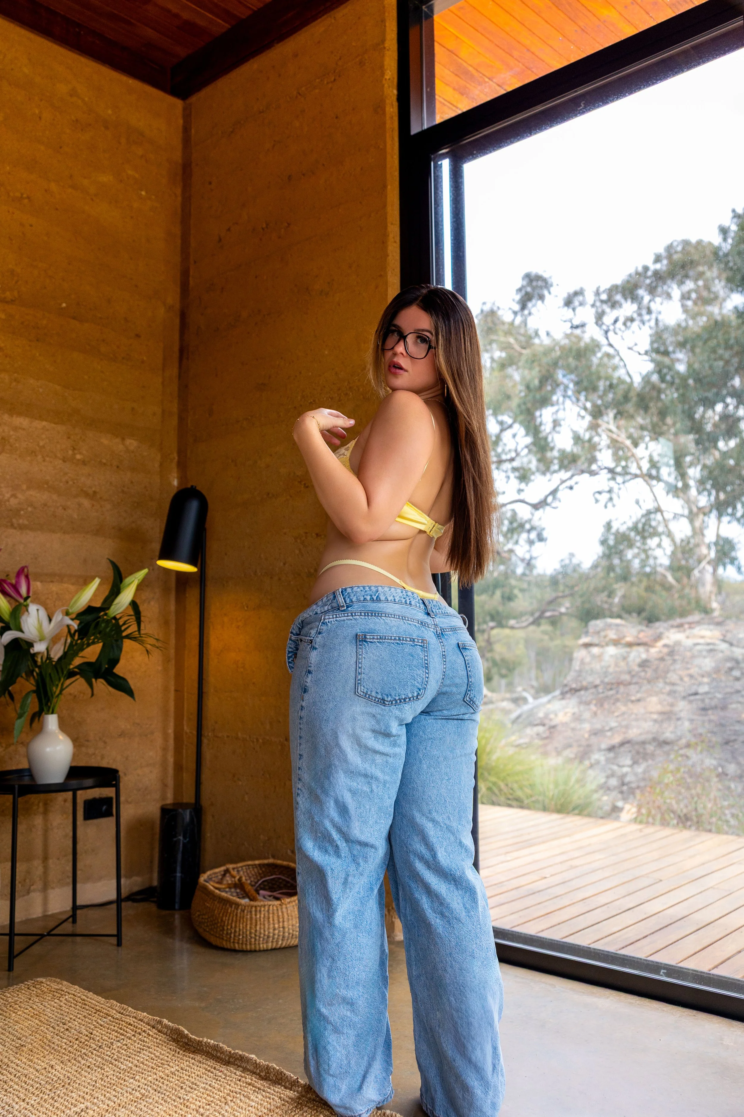 A woman with glasses wearing yellow lingerie and blue jeans looking over her shoulder indoors near a large window with a scenic outdoor view.