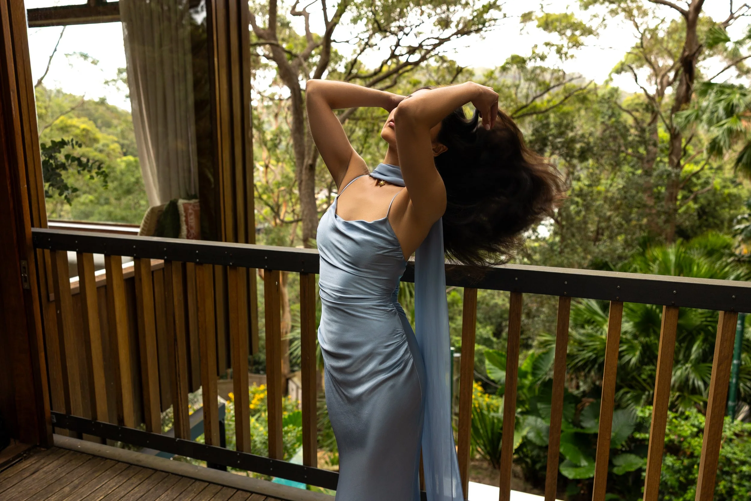 A woman dressed in a light blue satin dress standing on a wooden deck, with a natural green forest in the background, her head tilted back and arms raised, touching her hair.