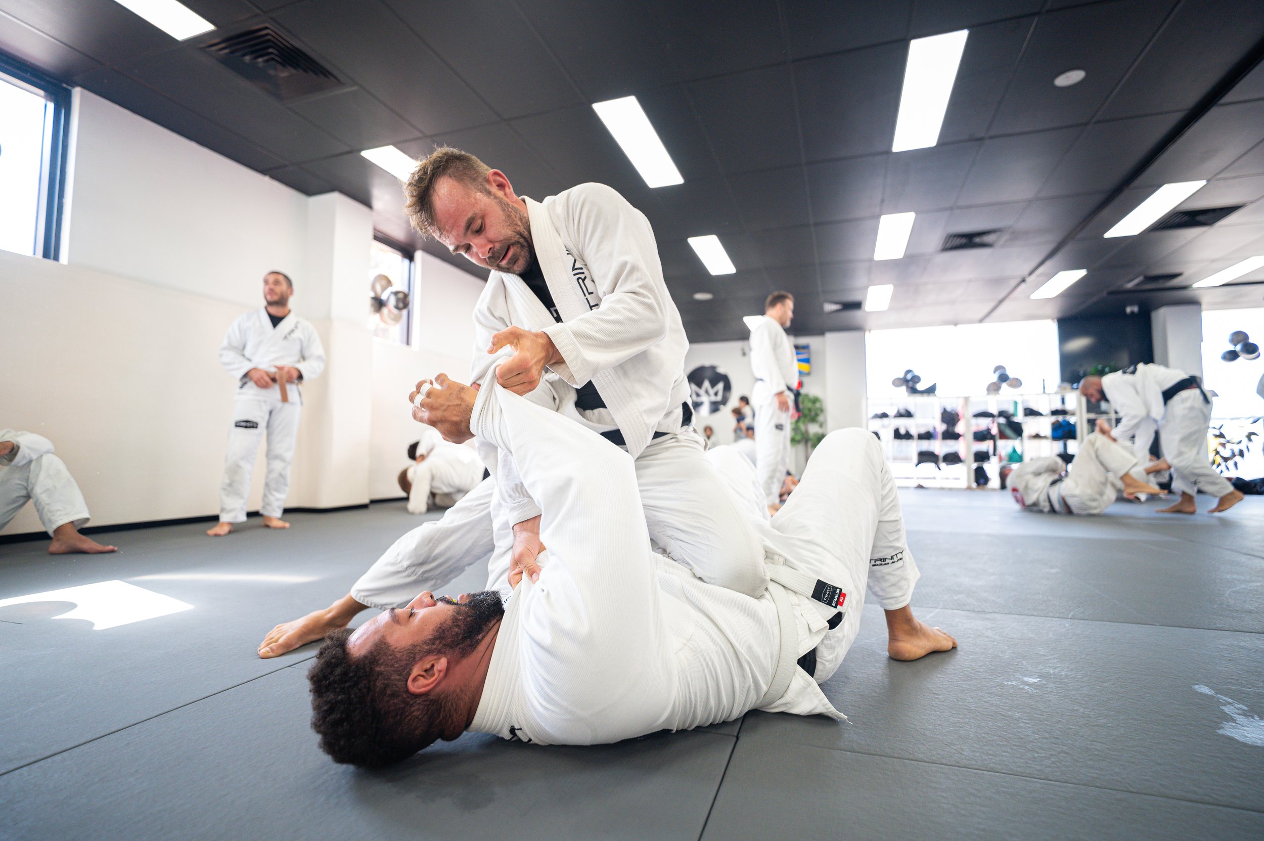 Brazilian Jiu-Jitsu training session with multiple students in white gis practicing on mats, including one person helping another with a grappling technique.