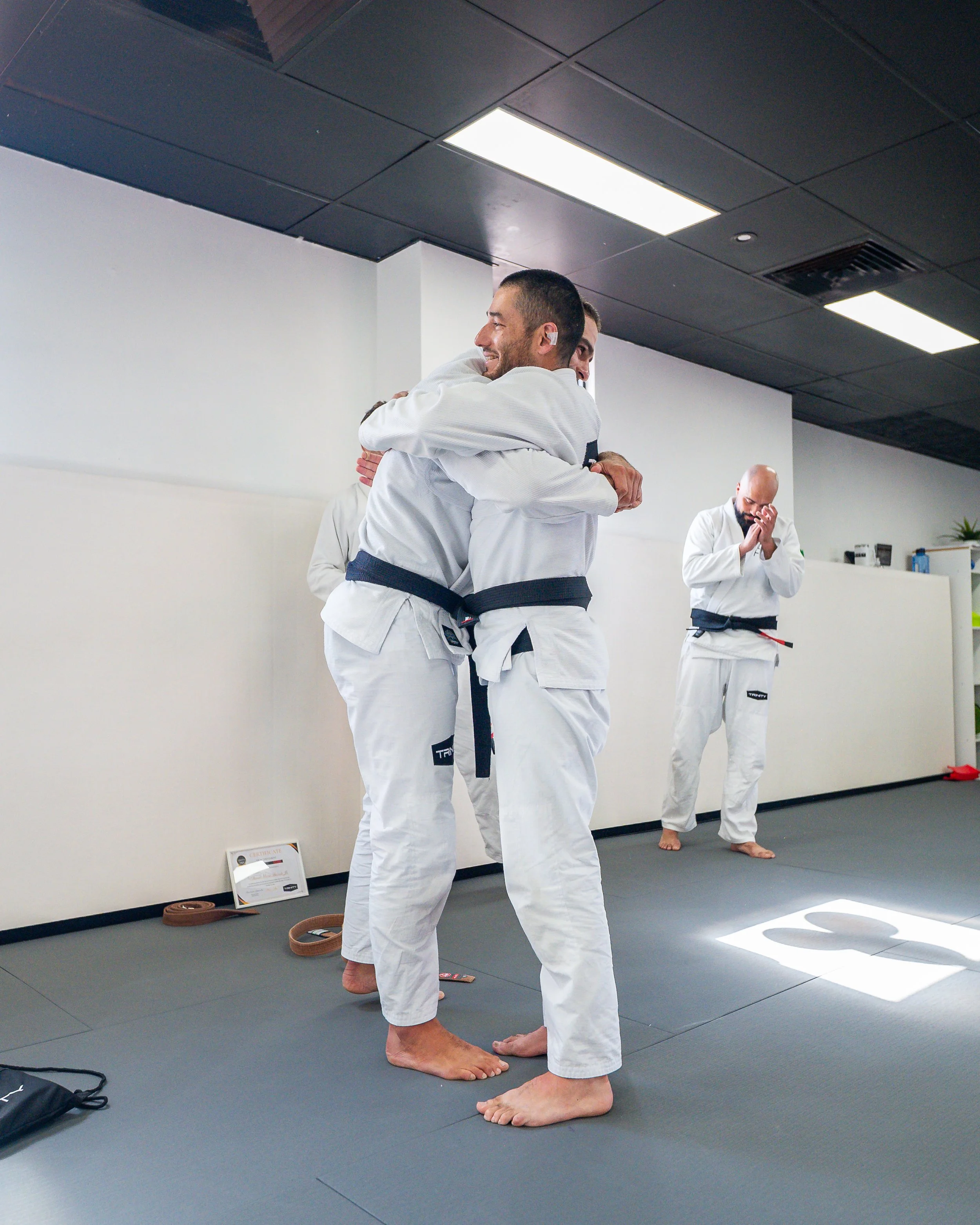 Two men in white martial arts uniforms hugging, with two other men in martial arts uniforms, one with hands clasped in front, in a martial arts dojo.