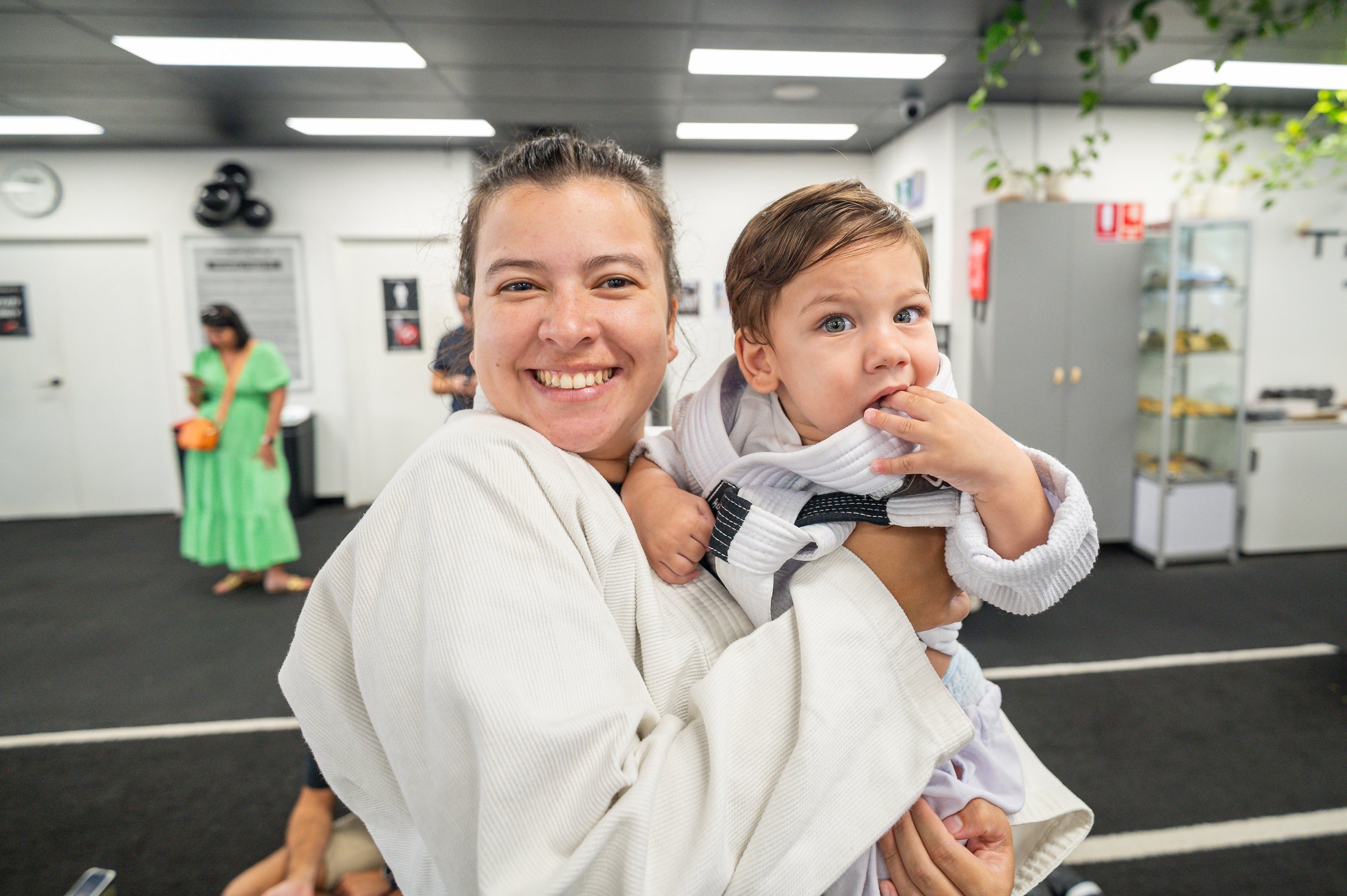 A woman in a white martial arts uniform smiling and holding a young boy in a white martial arts uniform inside a training facility, with other people and equipment in the background.