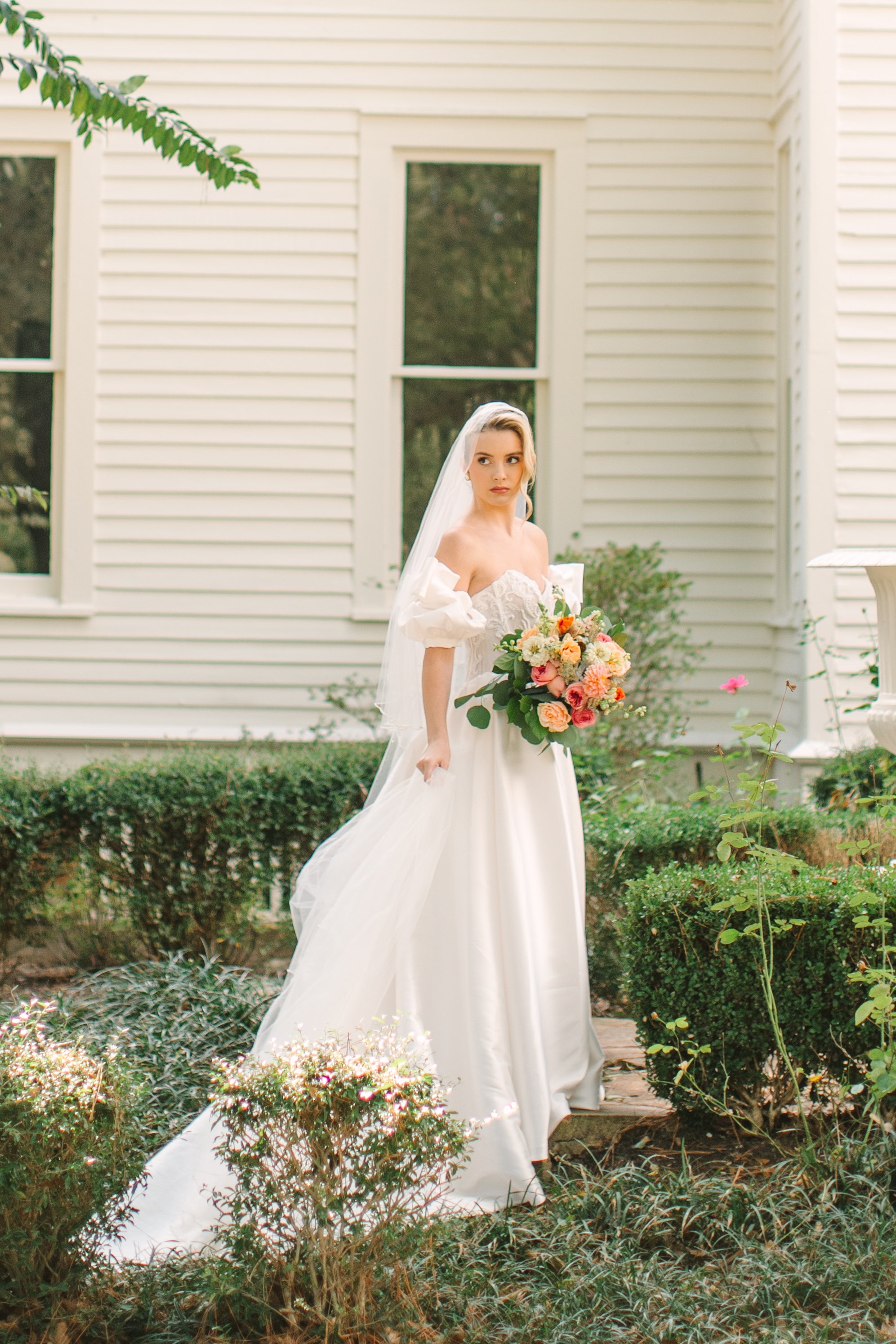 A bride in a white wedding dress with an off-shoulder design, holding a large bouquet of pink, peach, and white flowers, standing outdoors in front of a white house with green bushes and plants.