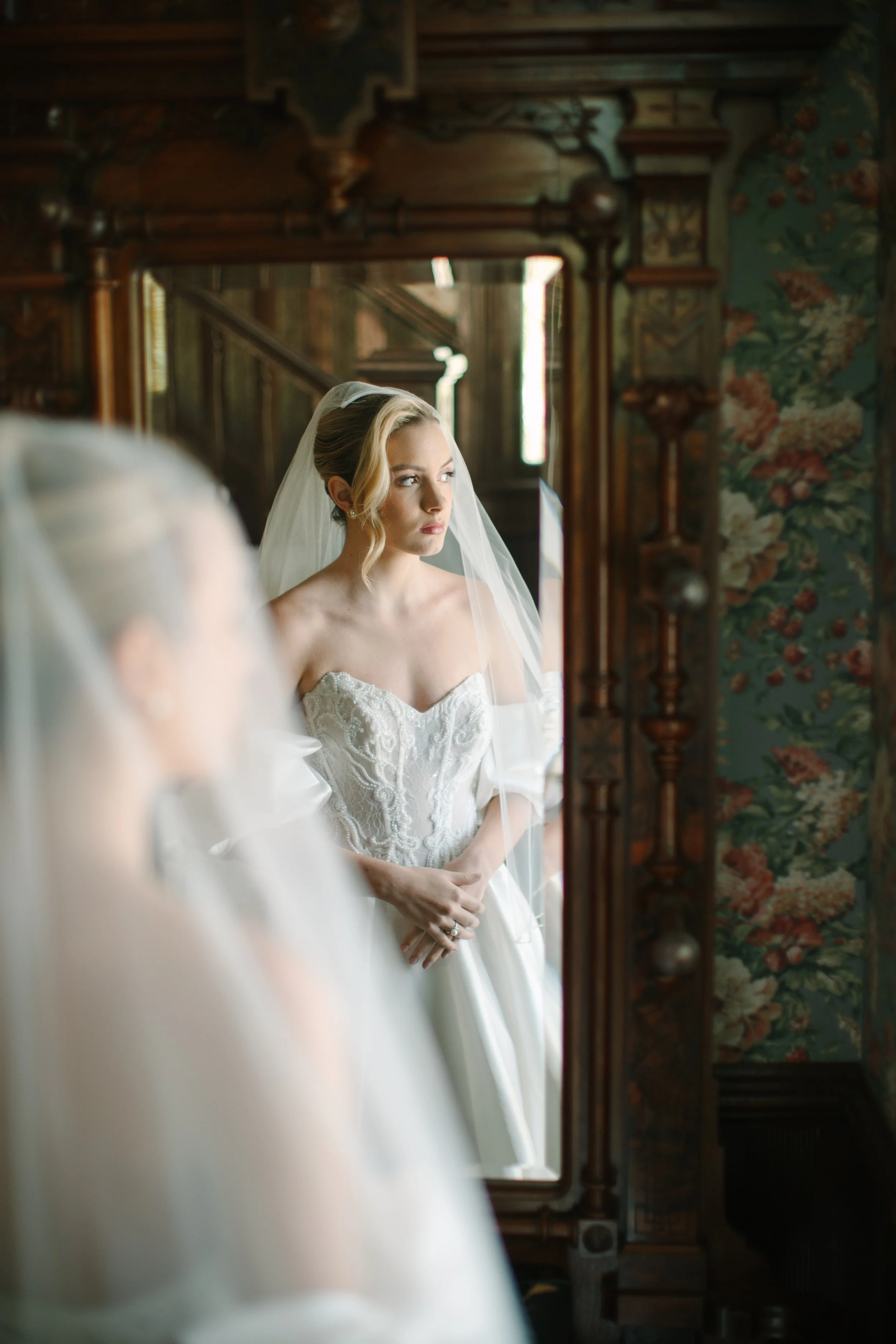 Bride looking at herself in an ornate wooden mirror, dressed in a white wedding gown with lace details and a veil, standing in a vintage decorated room with floral wallpaper.