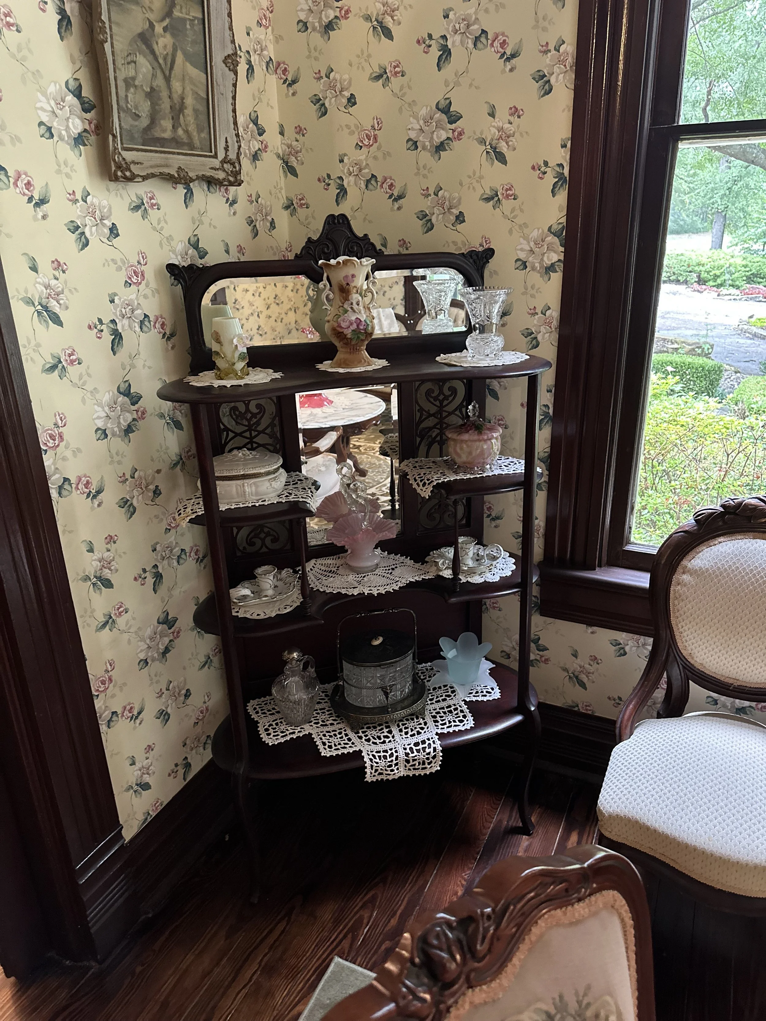 A wooden corner shelf with ornate detailing displays various glassware and decorative items, set against floral wallpaper next to a window with lush greenery outside.