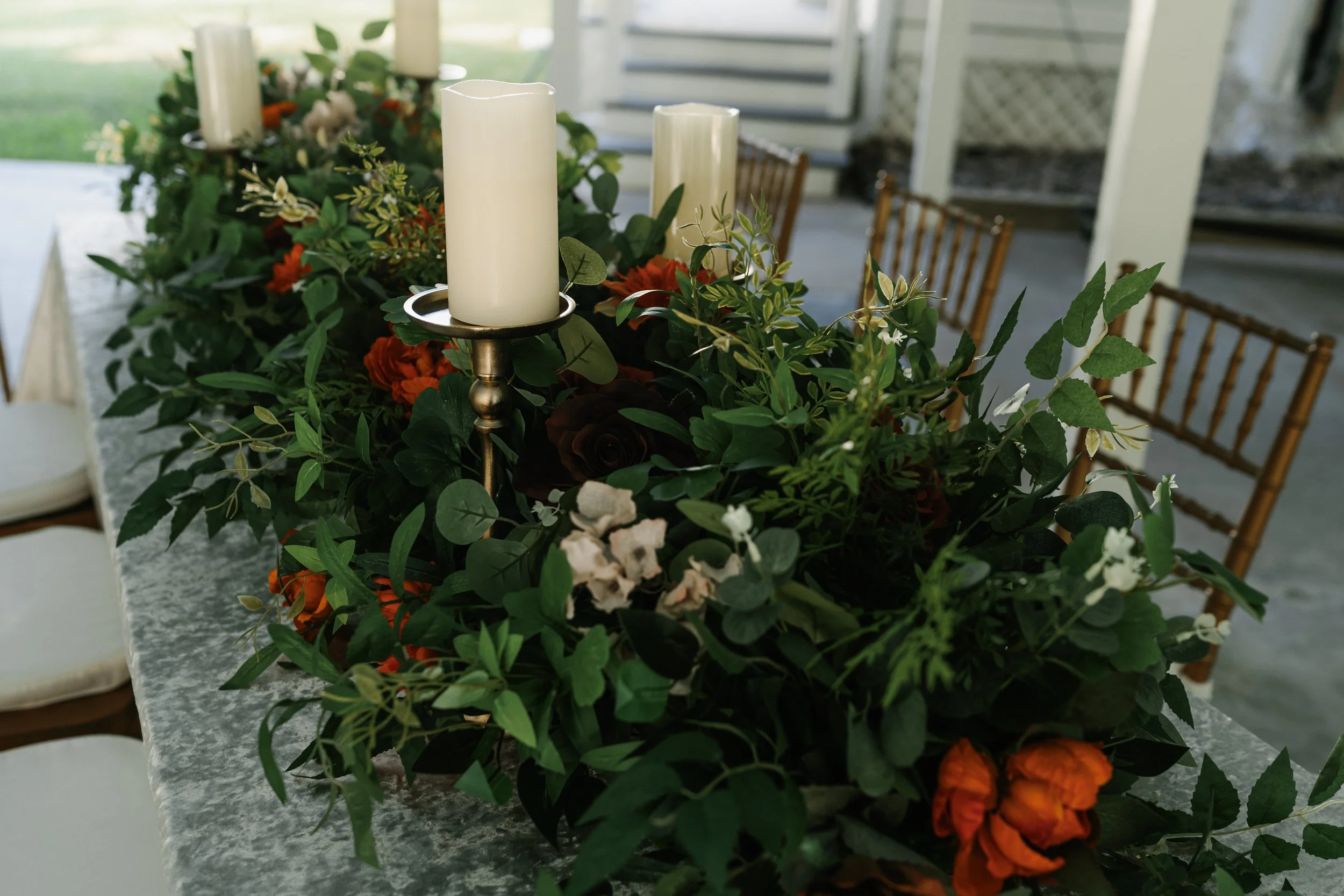 A long rectangular table decorated with a lush green floral centerpiece and white candles on candle holders, set in a bright indoor space.