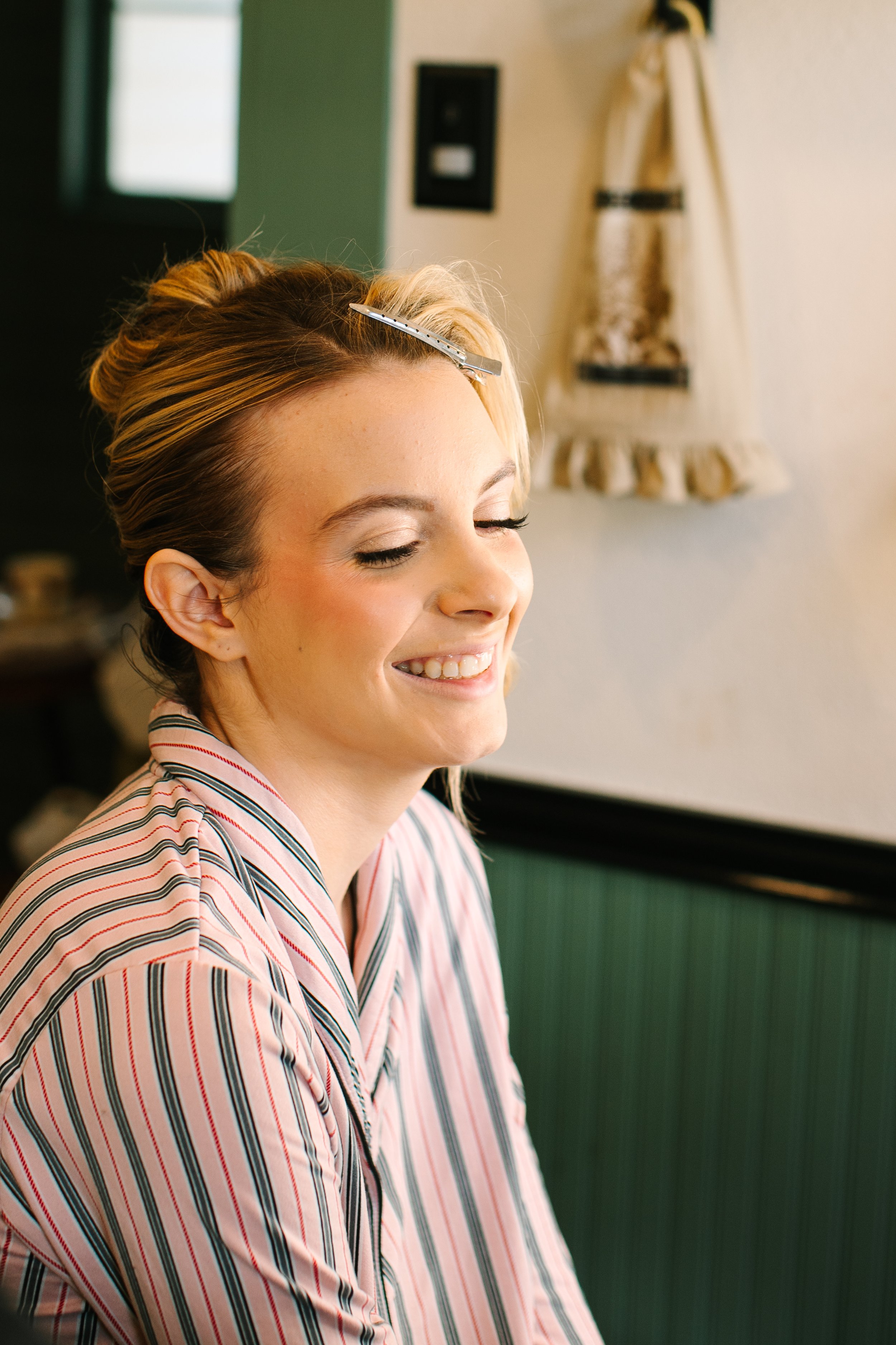 A woman with styled hair, a hair clip, and makeup smiling with her eyes closed, wearing a striped shirt in a cozy, decorated home.