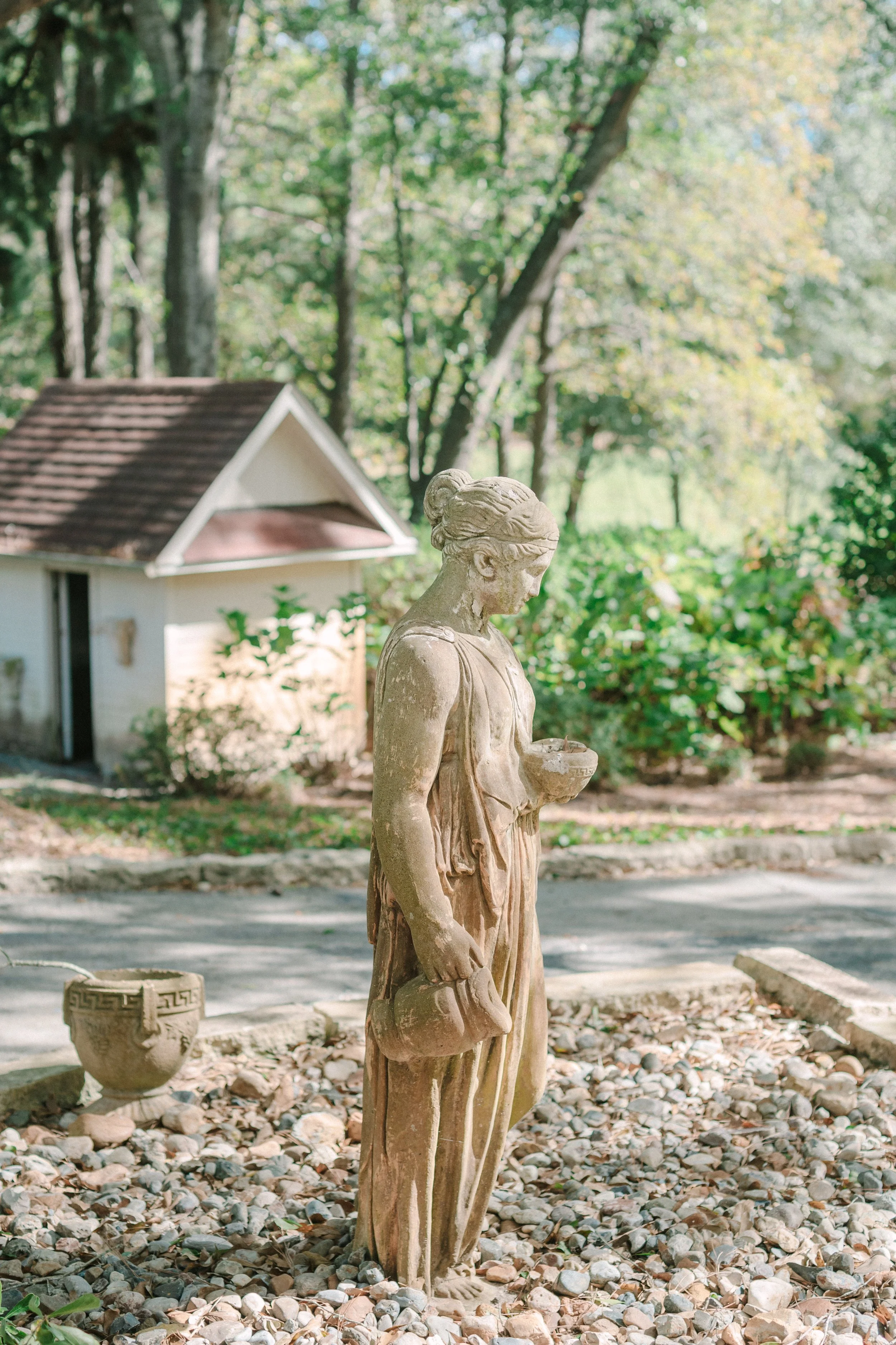 A stone statue of a woman holding a bowl and a watering can, standing on a bed of pebbles in a garden with a small building and trees in the background.