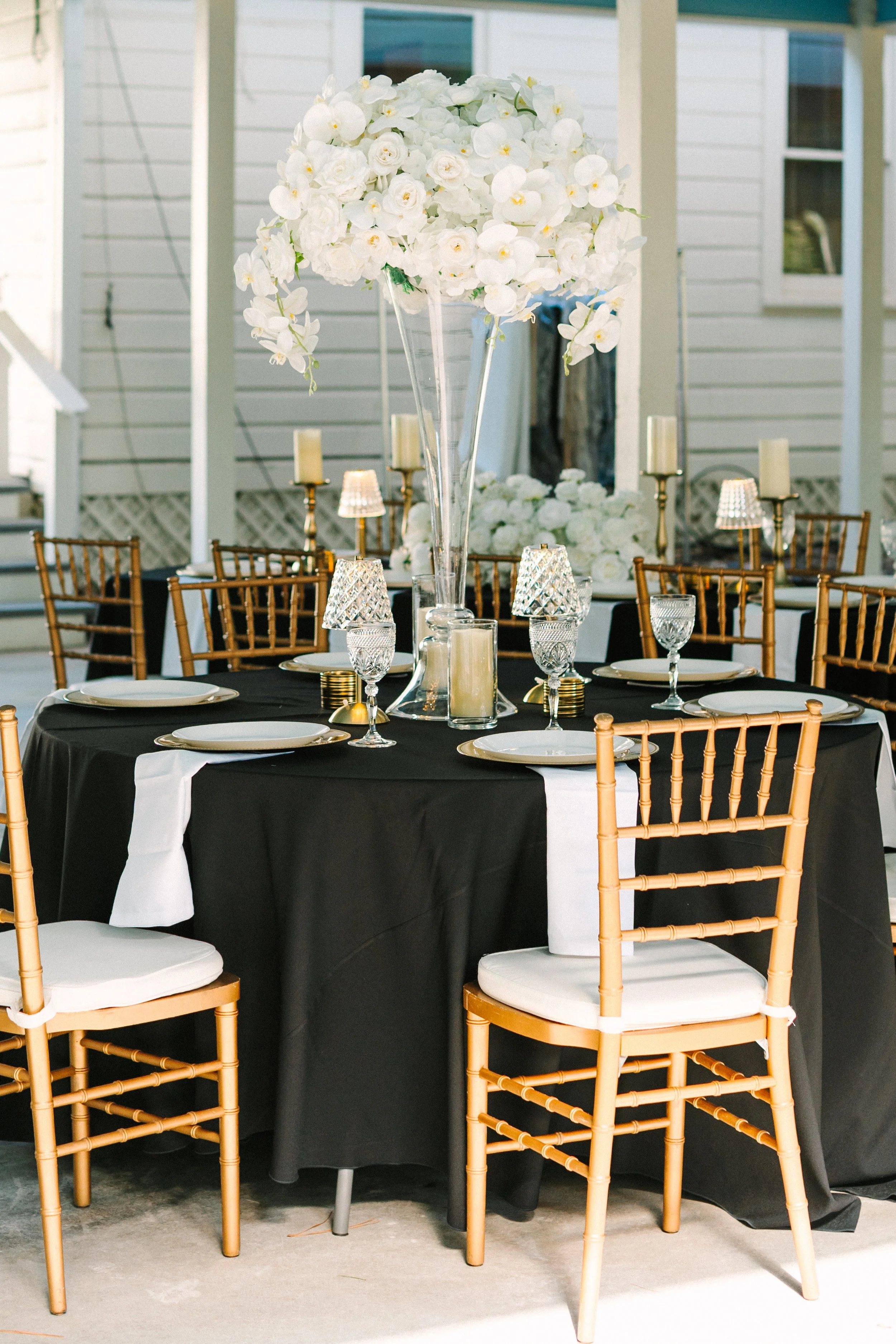Elegant outdoor table setting with black tablecloth, white plates, gold chairs, and a tall floral centerpiece of white flowers, surrounded by candles and crystal glasses.