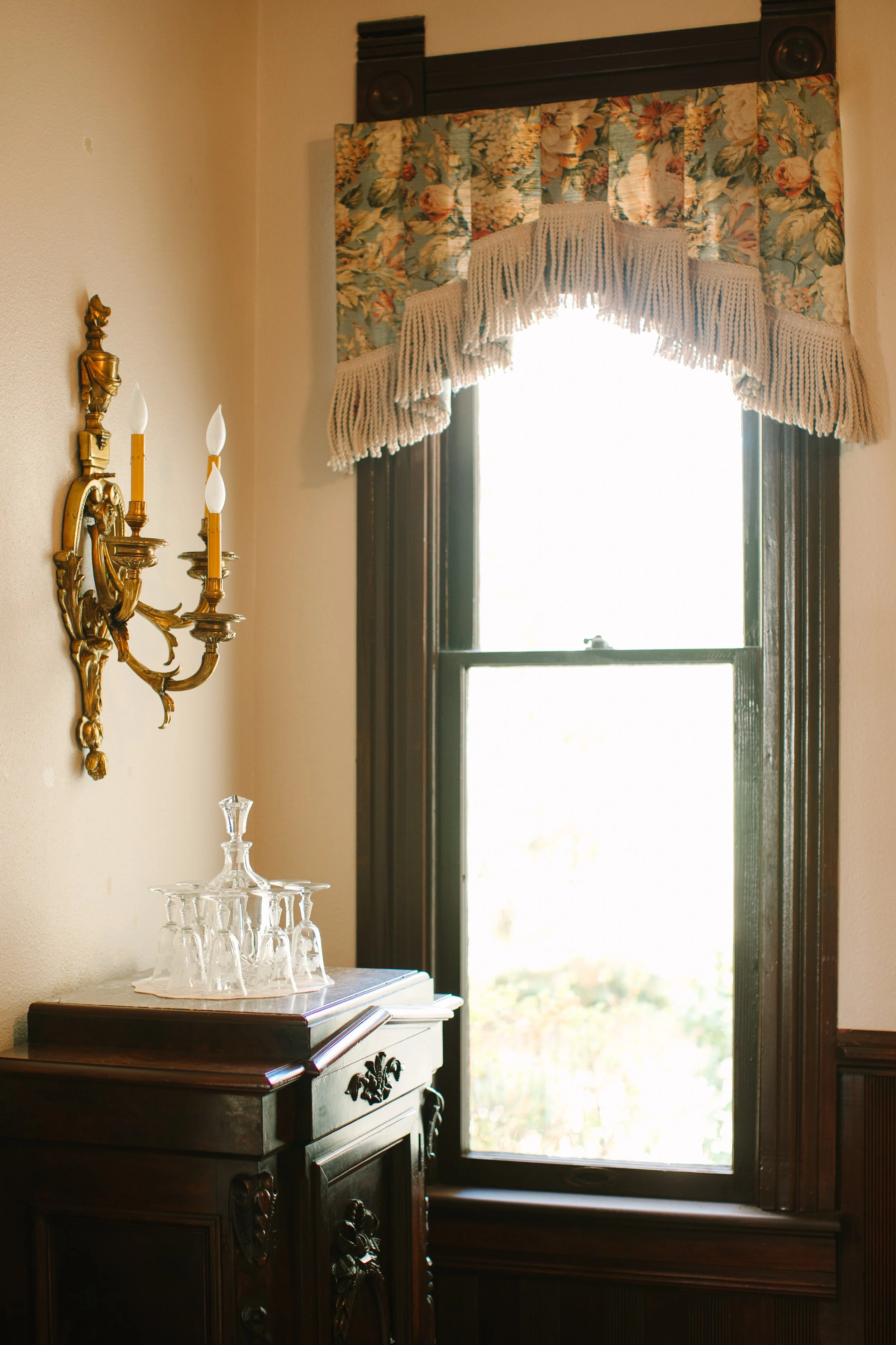 A vintage-style interior corner with a window covered by floral curtains with a scalloped lace valance. There is a dark wooden sideboard with a glass decanter and glasses on top, and a gold wall sconce with three candle-shaped lights on the beige wal