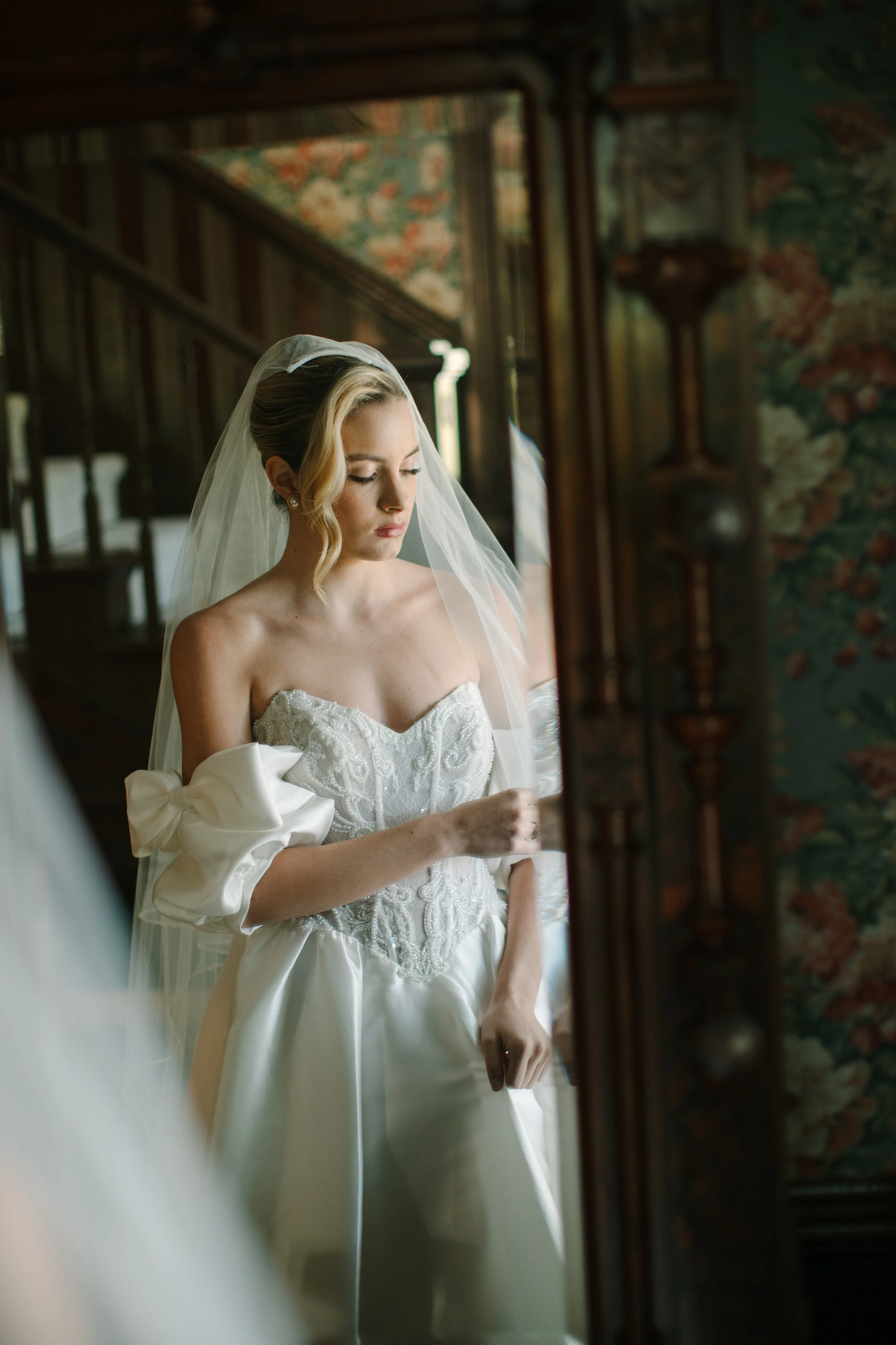 Young woman in a wedding dress with puffed sleeves and veil looking at herself in a mirror.