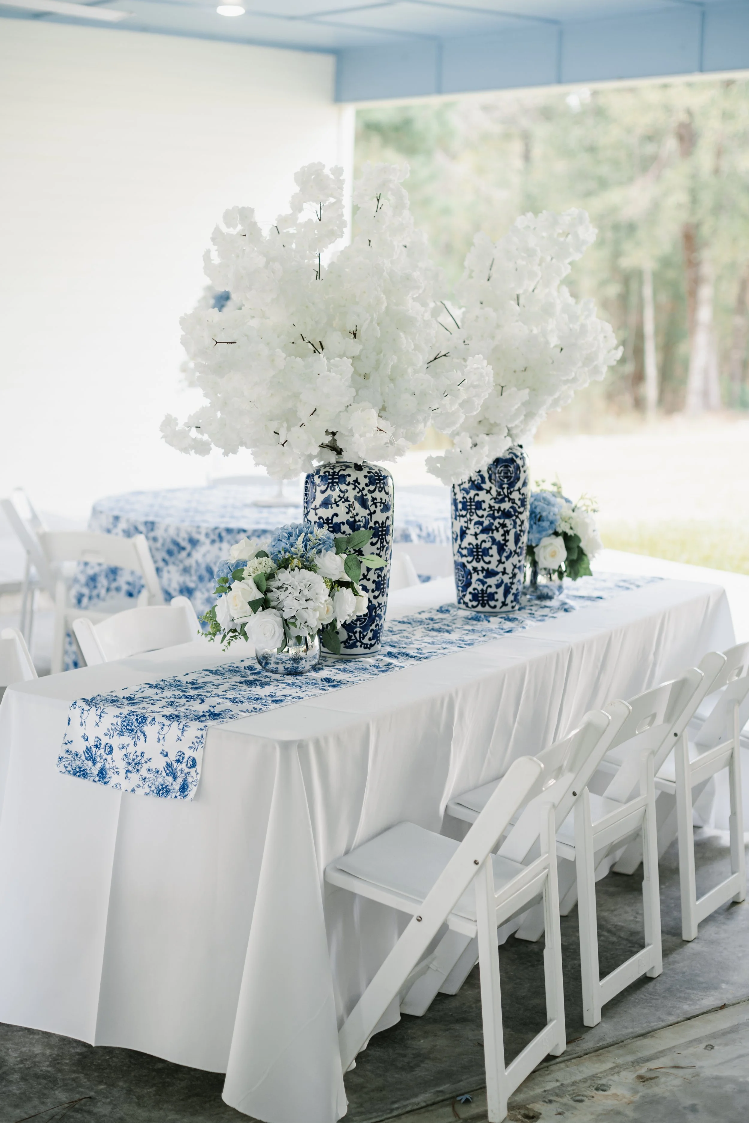 A wedding or event table decorated with blue and white floral arrangements, featuring large white flowers in blue-patterned vases and smaller white and blue flowers in a crystal vase, on a white tablecloth with a blue floral runner, set up in a brigh