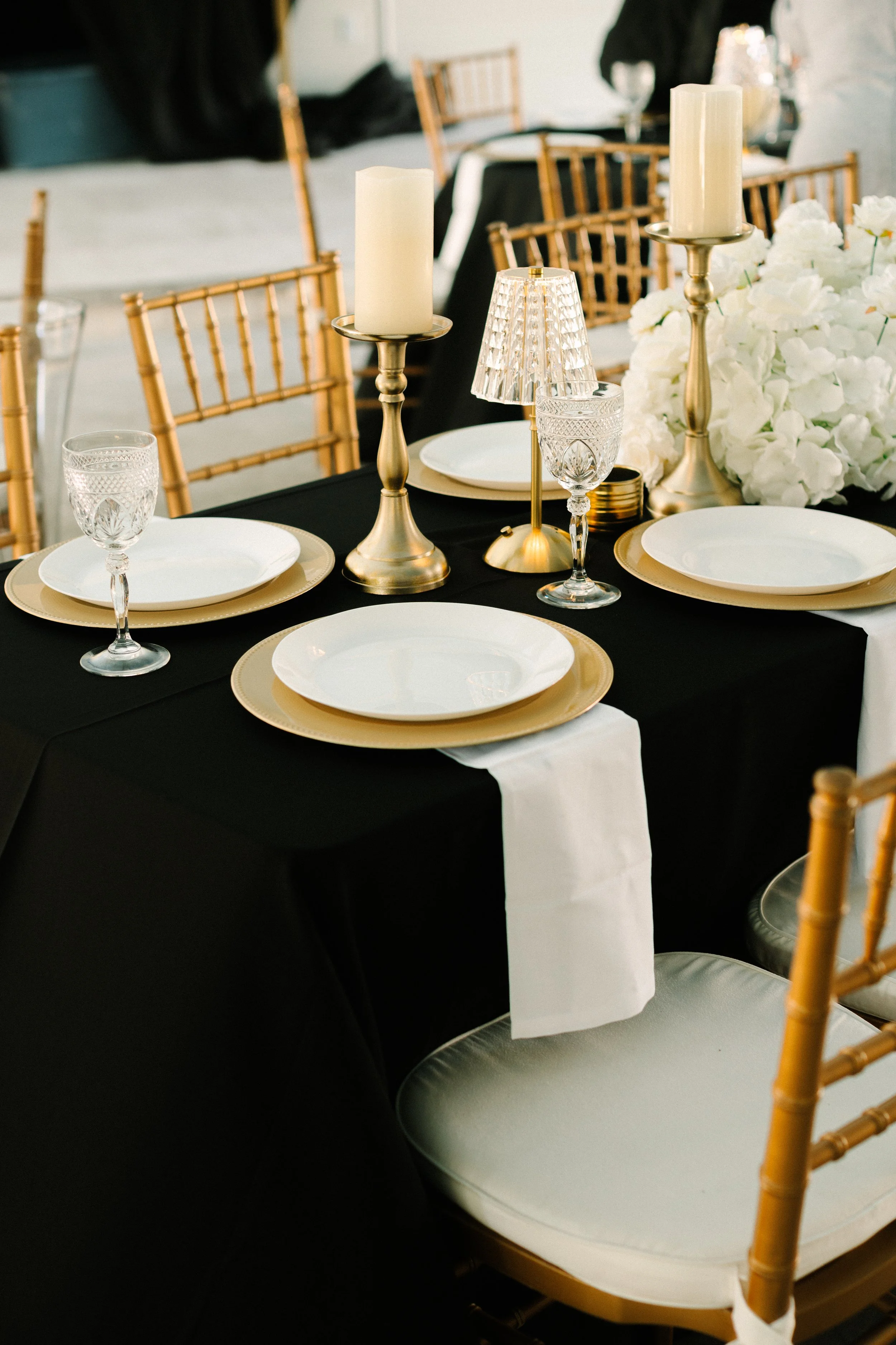 A formal dining table decorated with gold charger plates, white dinner plates, crystal glasses, tall gold candlesticks with white candles, a crystal table lamp, and a large white floral arrangement, set with a black tablecloth and gold chairs.