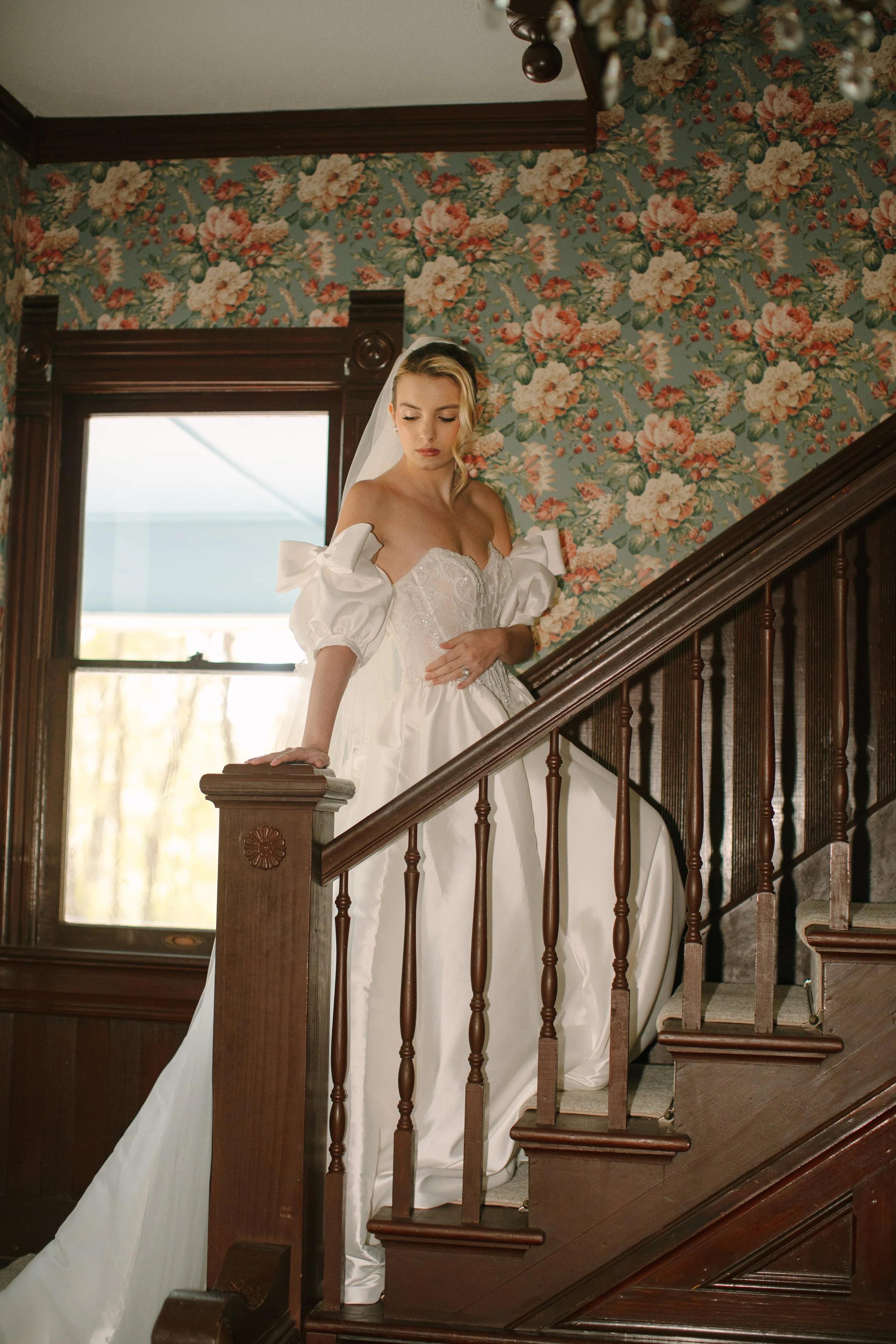 A bride in a white wedding gown with puffed sleeves, standing on a wooden staircase with floral wallpaper in the background.