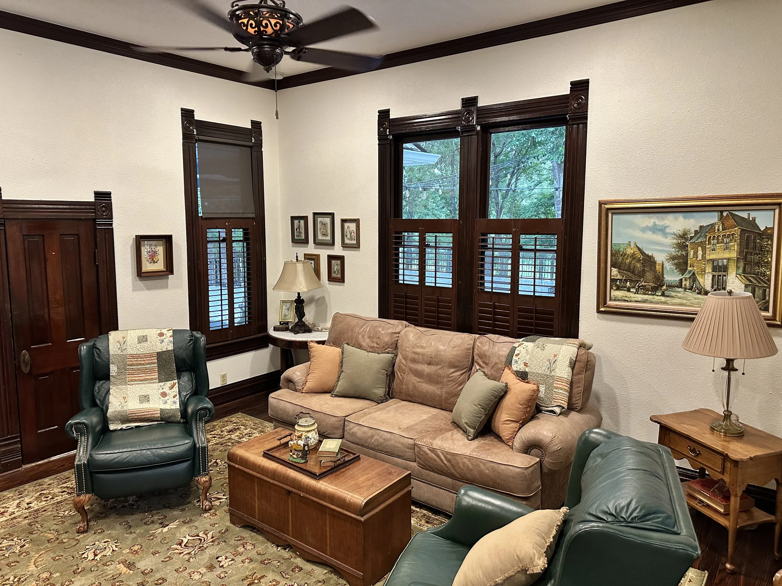 Living room with beige sofa, green armchair, and wooden coffee table, decorated with pillows and quilts, surrounded by hardwood floors, dark wood window frames, and framed wall art, with a ceiling fan overhead.