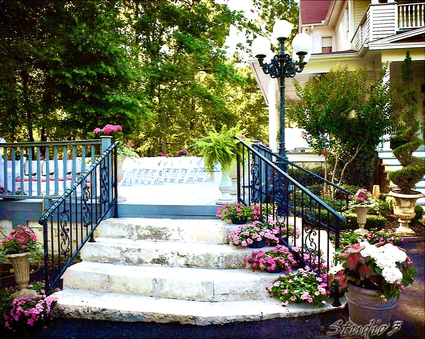 Exterior view of a house with stone steps, black metal railings, and colorful flowers in pots. There is a vintage-style lamp post near the steps, and the background features lush green trees and a patio with white chairs and plants.