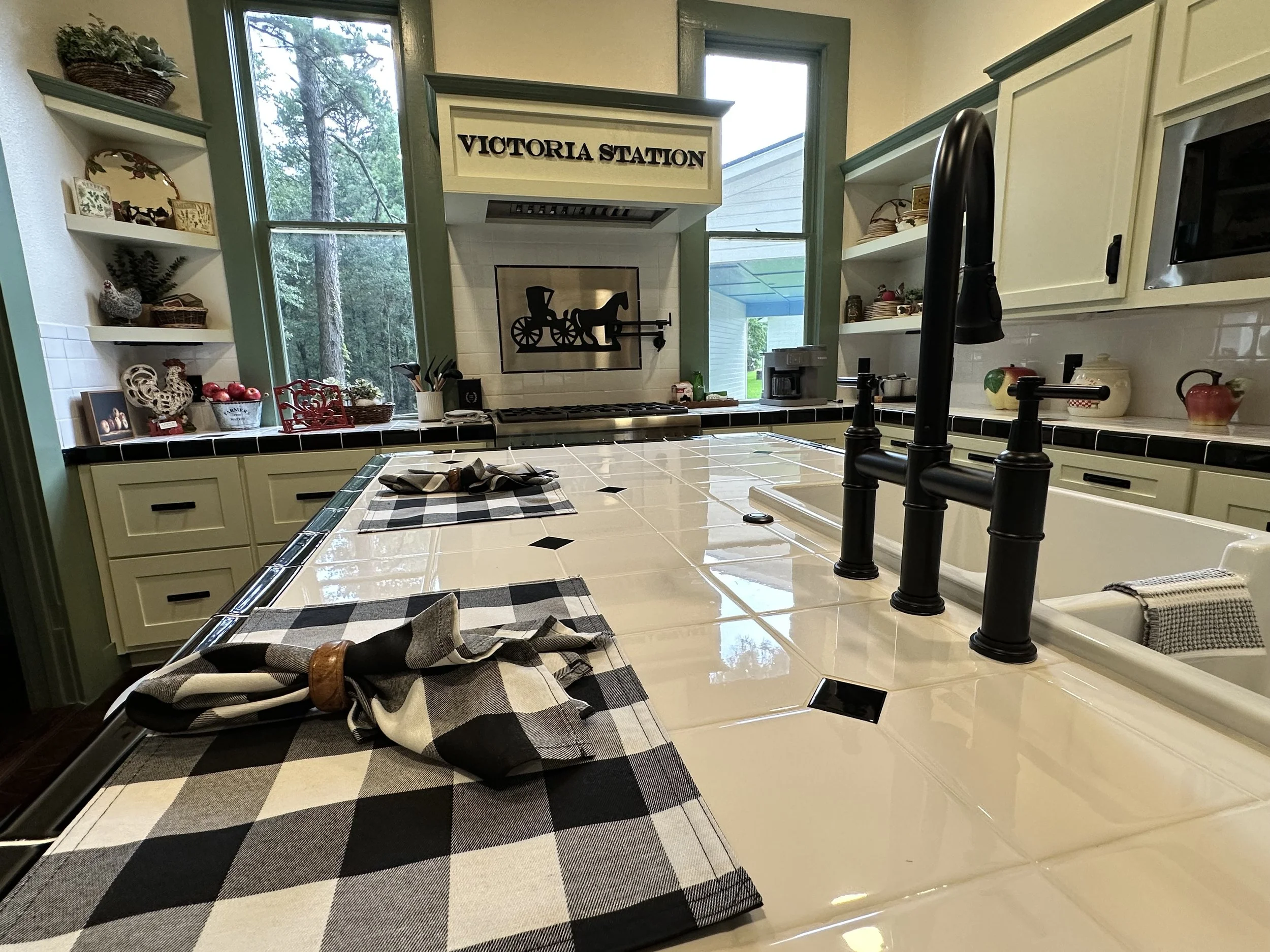 Kitchen with cream-colored cabinets, a large tiled island with a black faucet, black and white checkered dish towels, and shelves with various decorative items. A black-and-white wall art with a horse-drawn carriage, and a window showing a green outd
