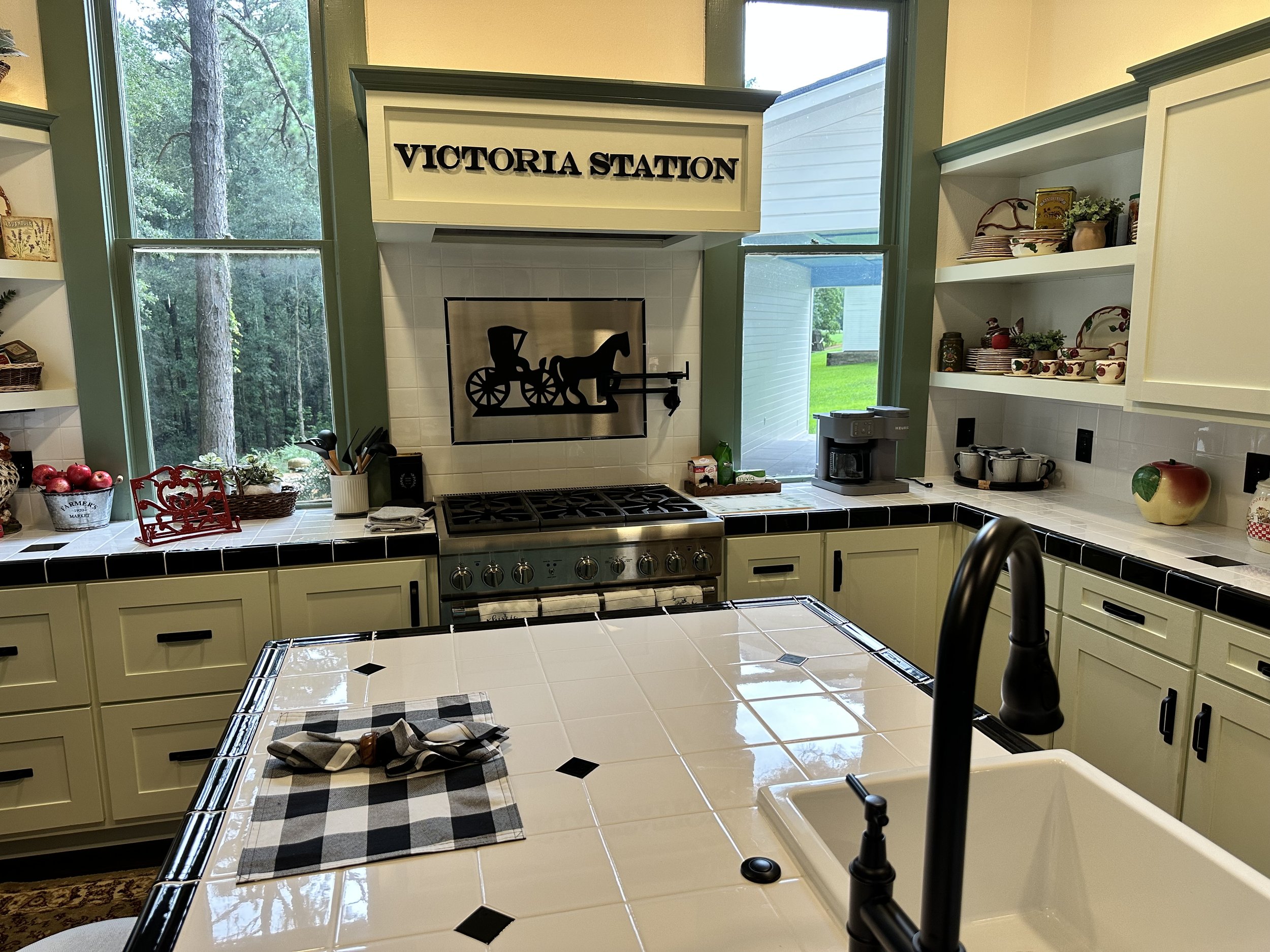 Kitchen with white cabinets, black countertop tiles, window with view of trees, and a sign reading 'Victoria Station' above a stove.
