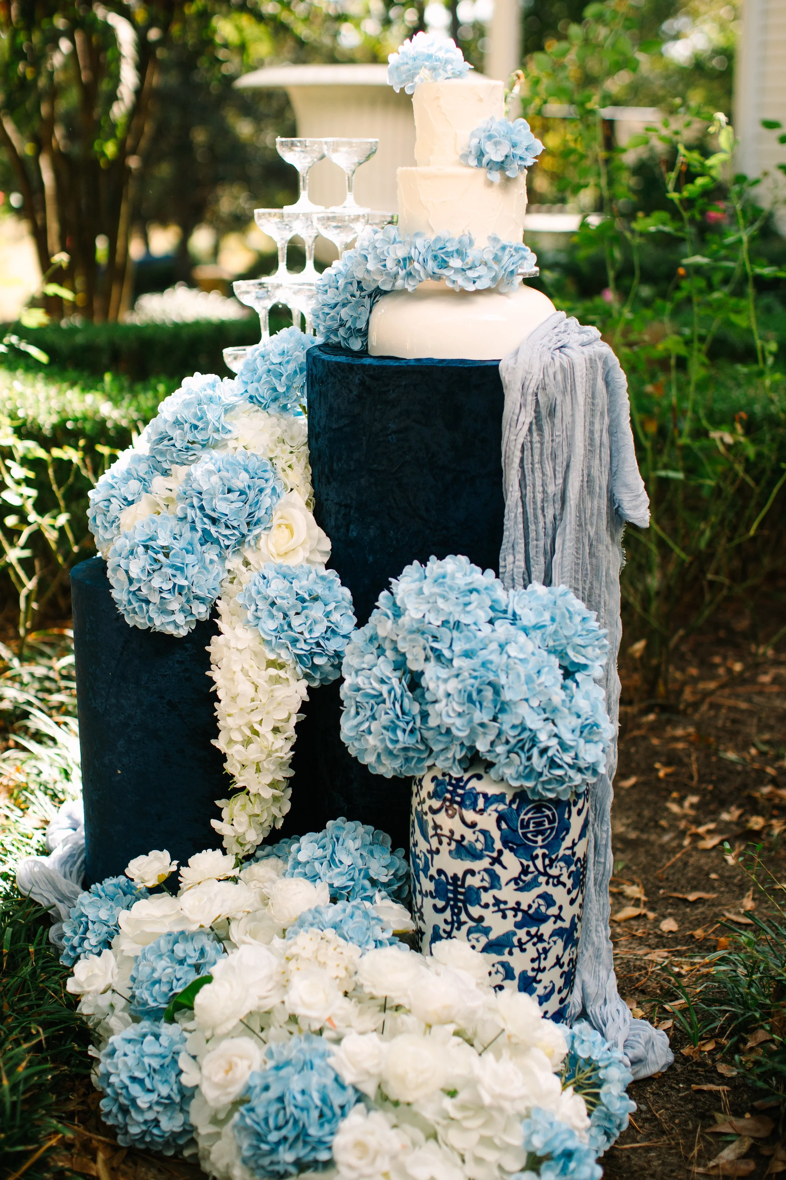 Elegant outdoor floral display with a tiered wedding cake, blue hydrangeas, white roses, and decorative vases, set in a garden.