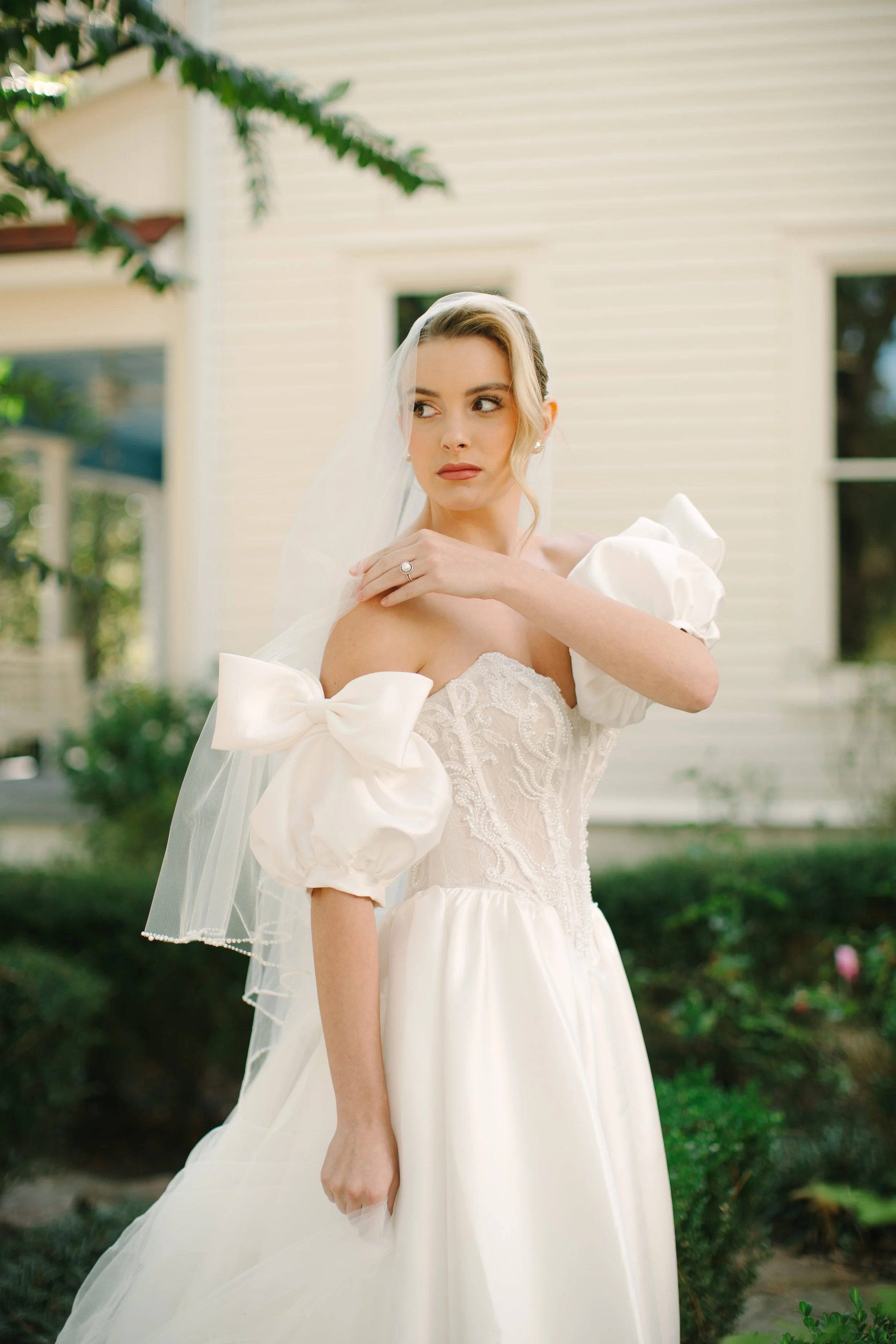A woman in a wedding dress with puffed sleeves and bows, standing outdoors with a veil over her head, in front of a house with beige siding and greenery.