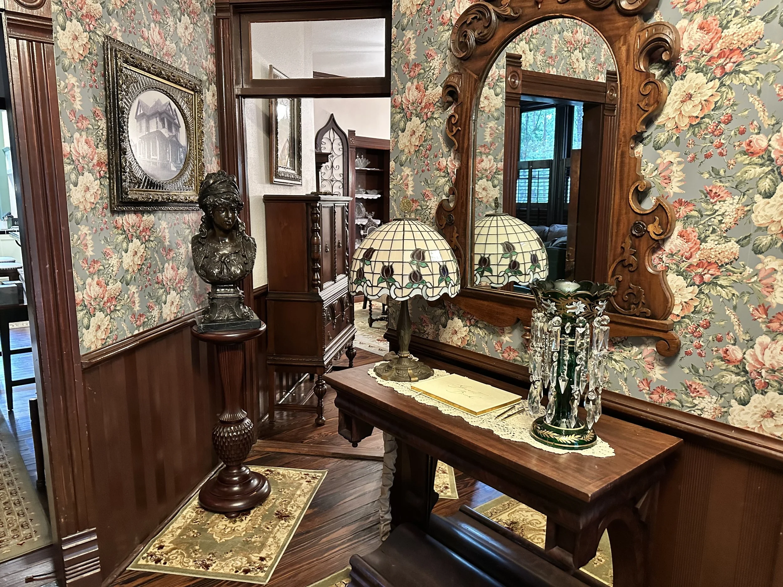 An antique wooden dresser with a large carved wooden mirror above it, decorated with a vintage stained glass lamp, a flower arrangement, and a bust sculpture of a woman. The room has floral wallpaper, wood-paneled lower walls, and wood flooring with 