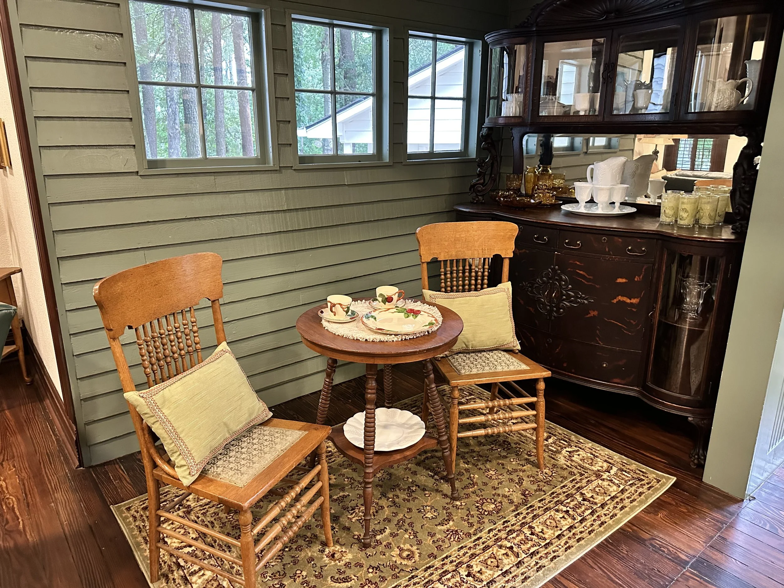A vintage dining nook with a small round wooden table, two wooden chairs with cushions, an ornate dark wood china cabinet filled with dishware, and a window showing a wooded outdoor scene.