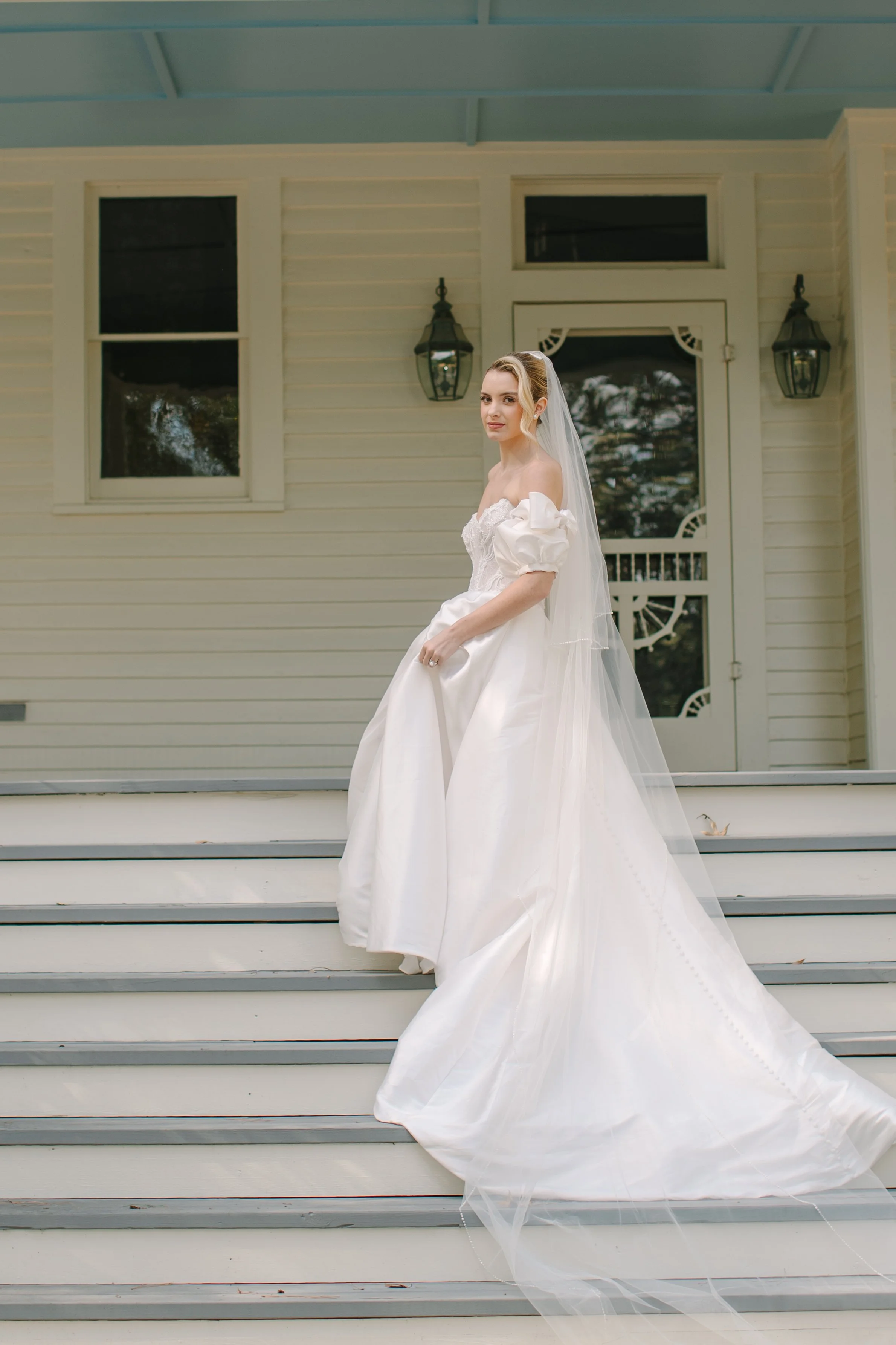 A bride in a white wedding dress with a long veil standing on the stairs outside a house with white siding and dark window panes.