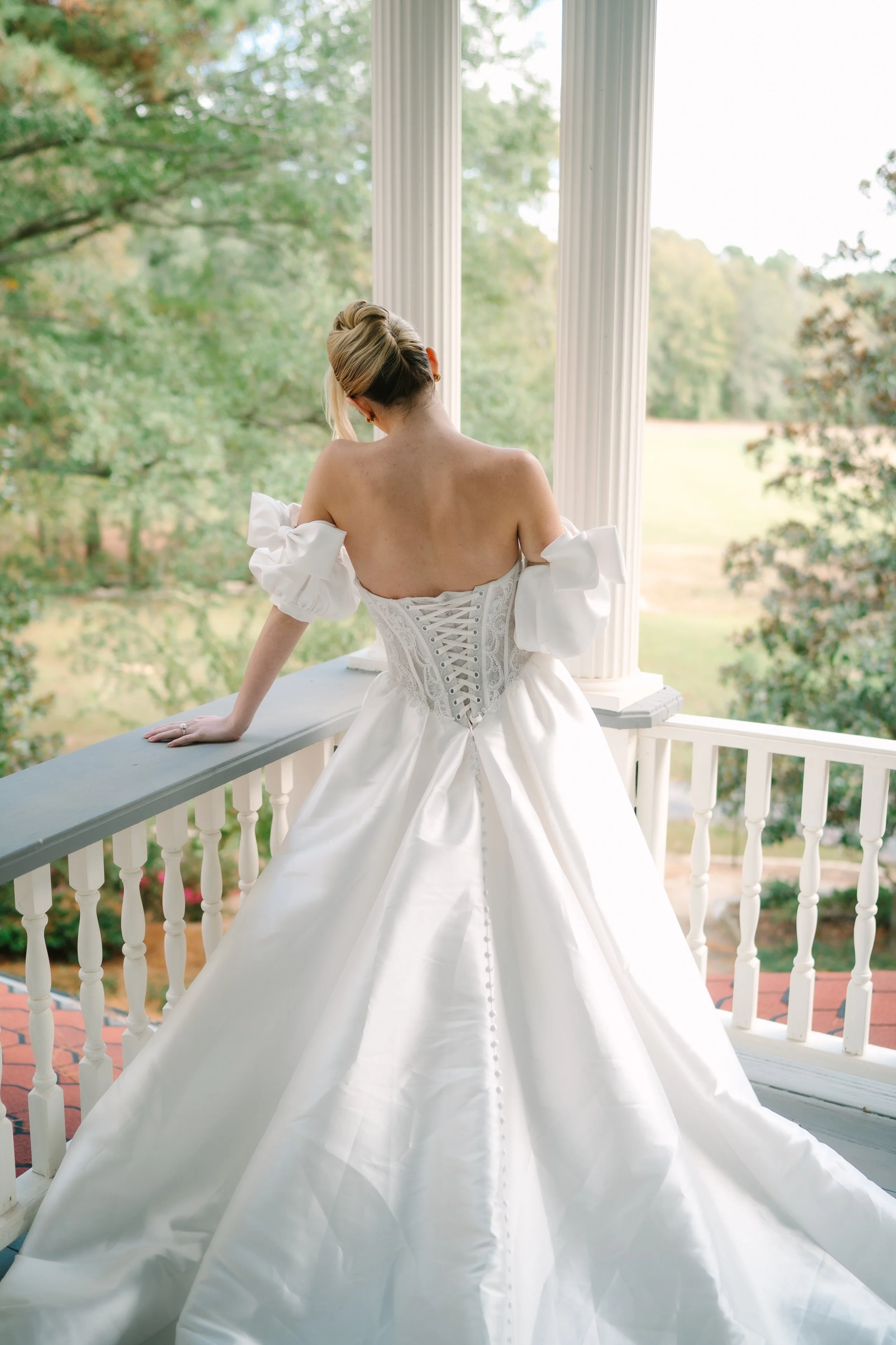 A woman in a wedding dress standing on a porch, looking out at greenery and trees.