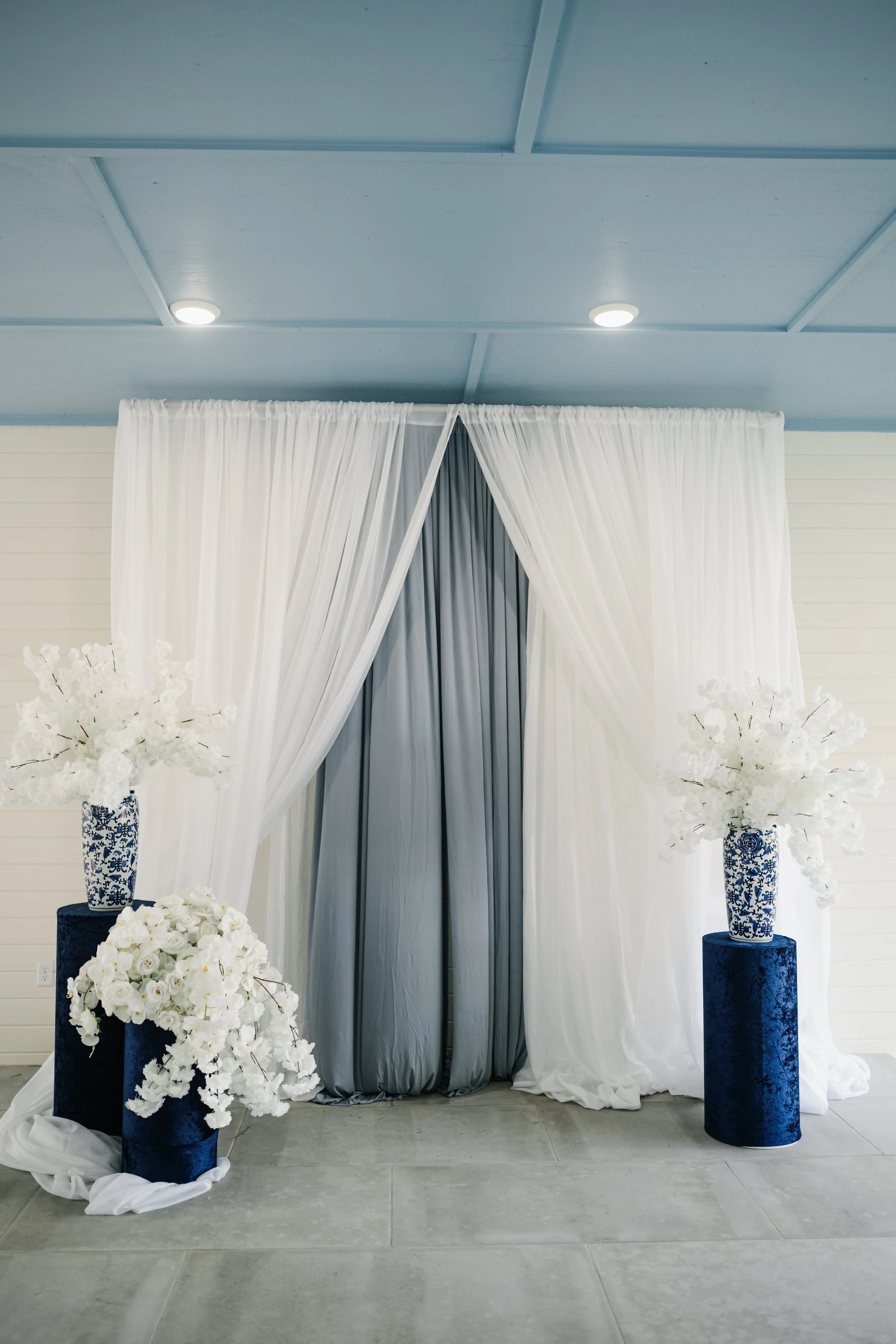 Elegant wedding backdrop with white and gray curtains, flanked by white floral arrangements in blue and white vases on blue pedestals.