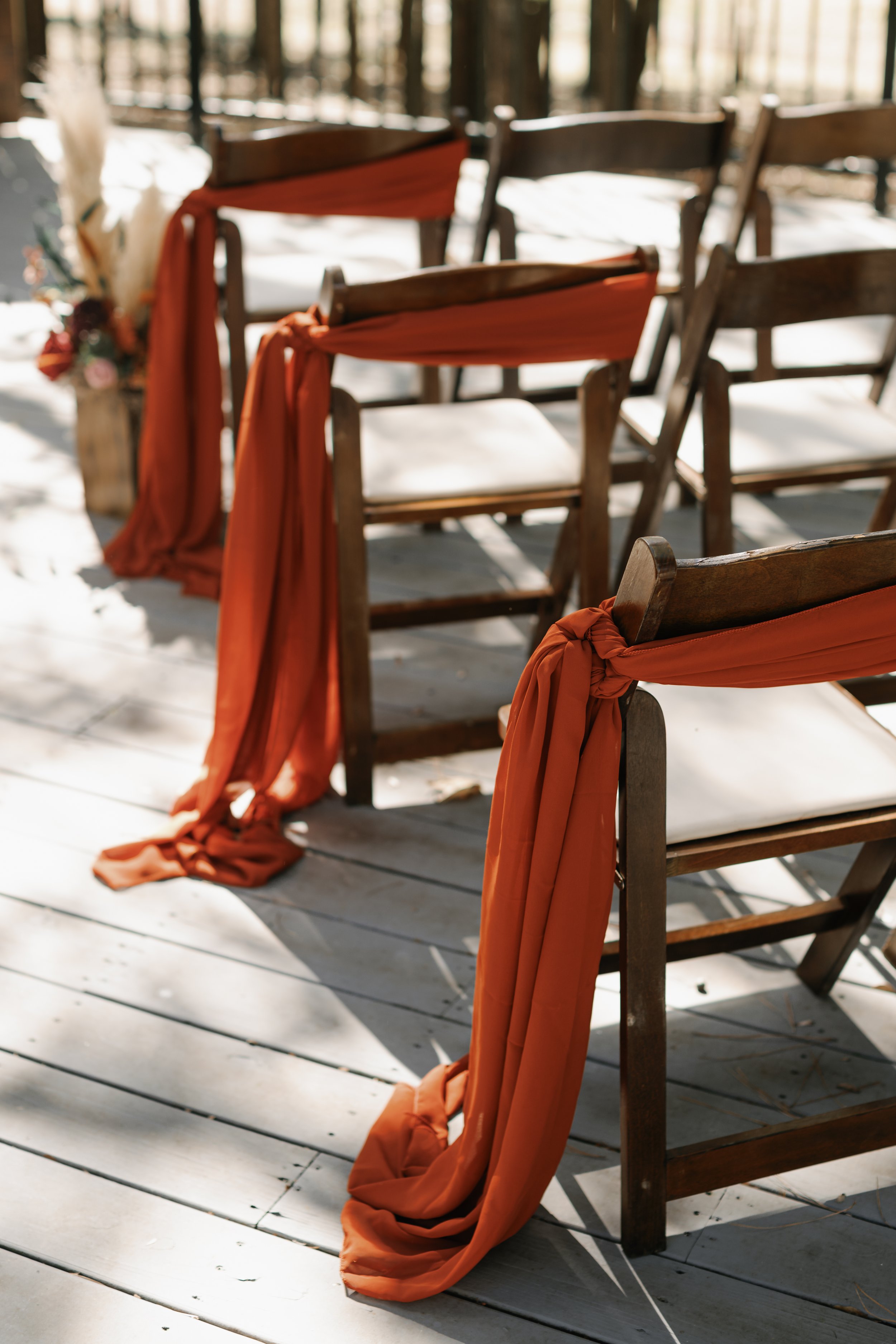 Wooden chairs decorated with rust-colored fabric and tied on the side, set up outdoors on a wooden deck with sunlight filtering through trees.