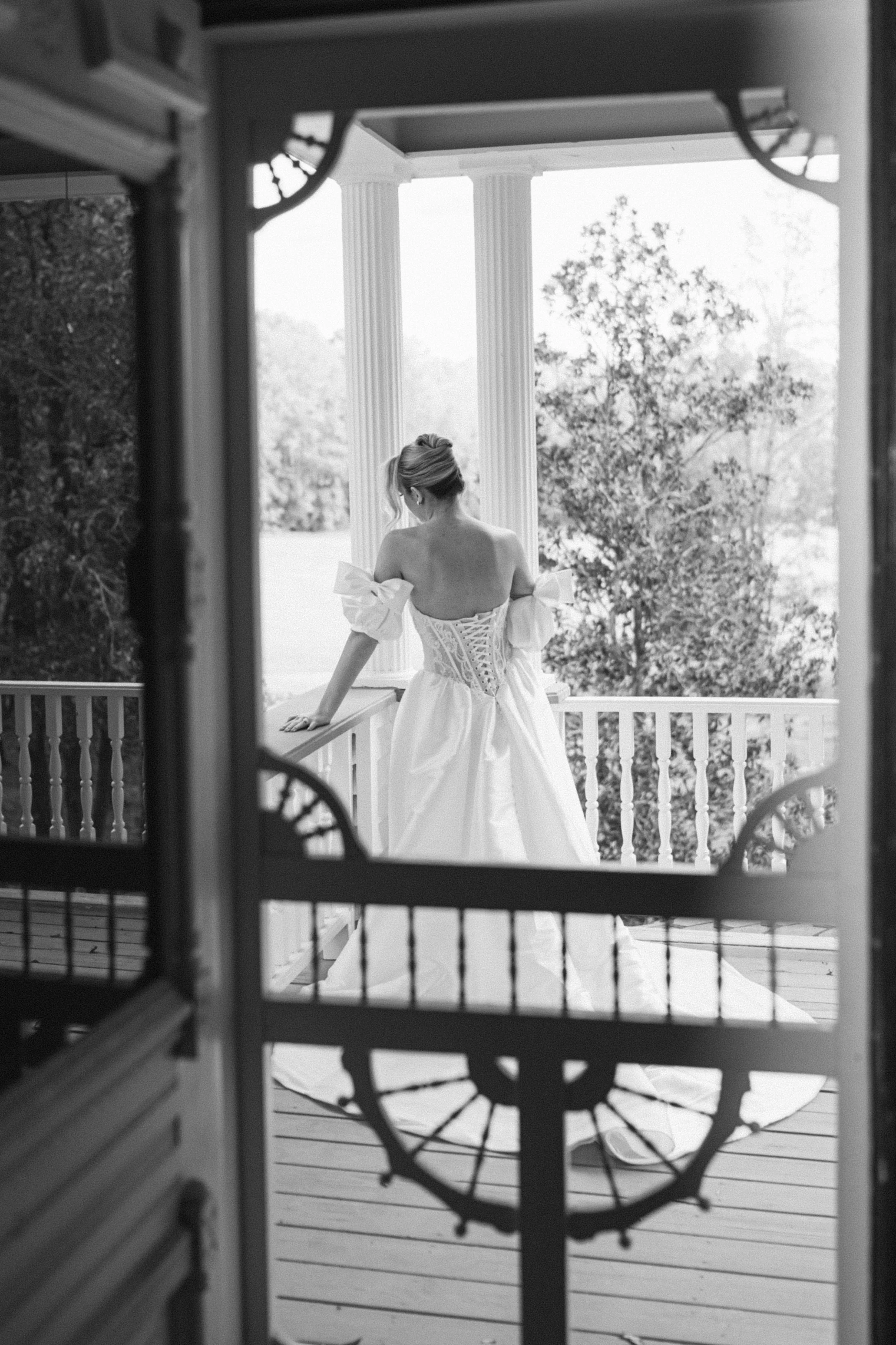A woman wearing a wedding dress standing on a porch, seen through a decorative window frame, with trees in the background.