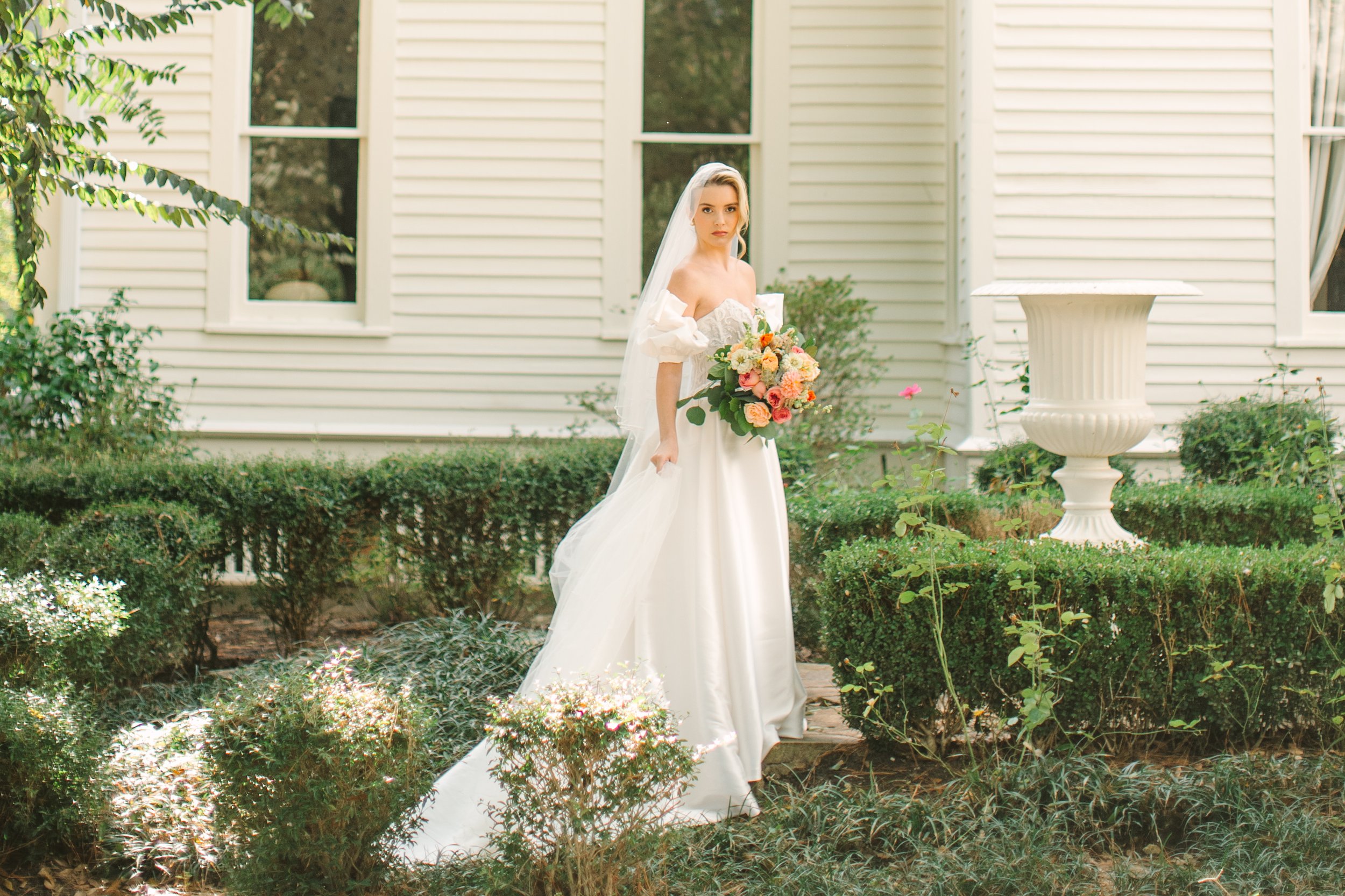 A bride in a white wedding dress with off-the-shoulder puff sleeves stands outdoors in front of a white house with shutters, holding a large bouquet of pink, peach, and white flowers, and looking at the camera.