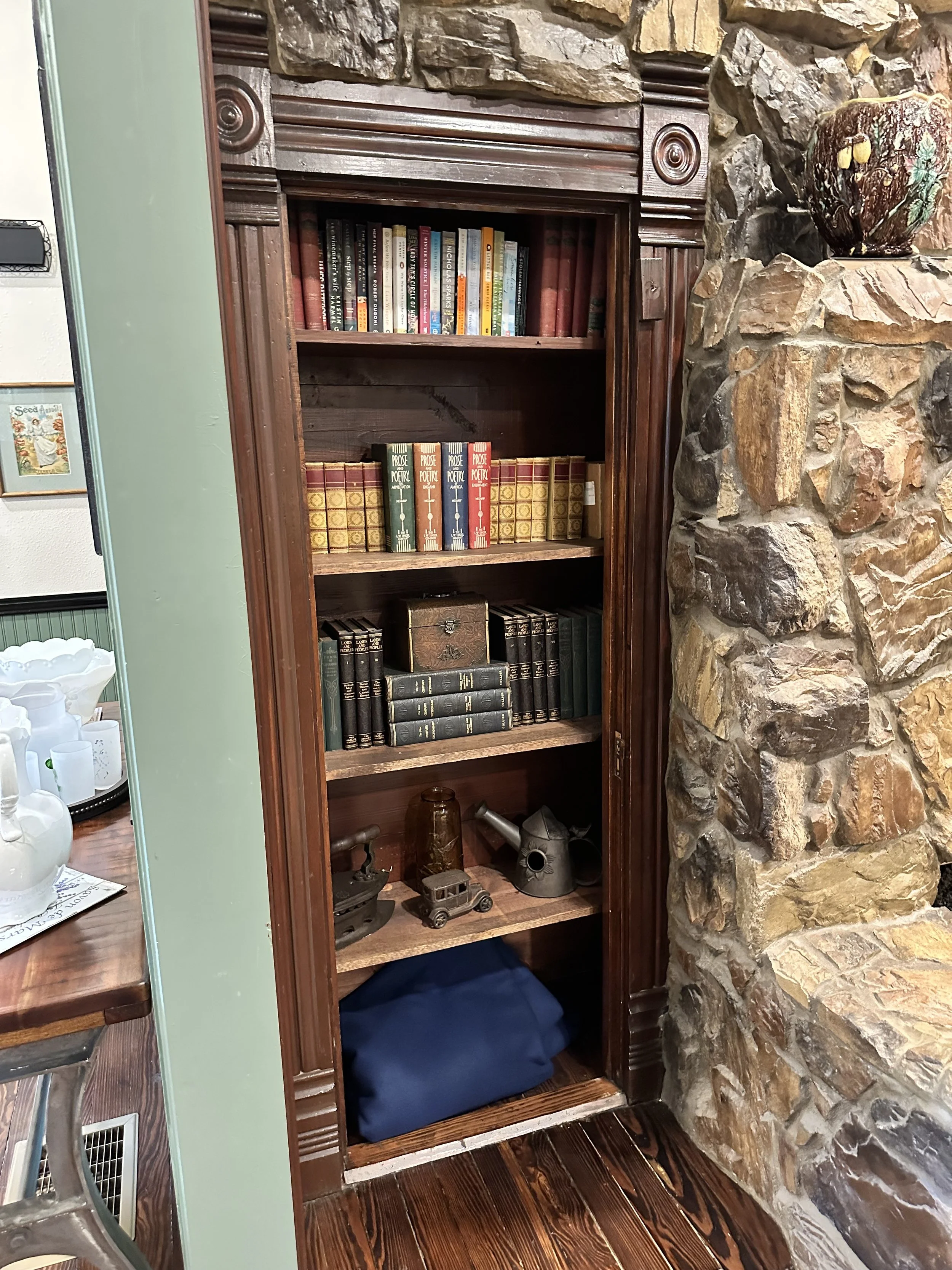 A tall wooden bookshelf with four shelves filled with books, located next to a stone wall with a decorative vase on top. The bottom shelf contains a blue bag and some small decorative items including antique toys and watering cans.