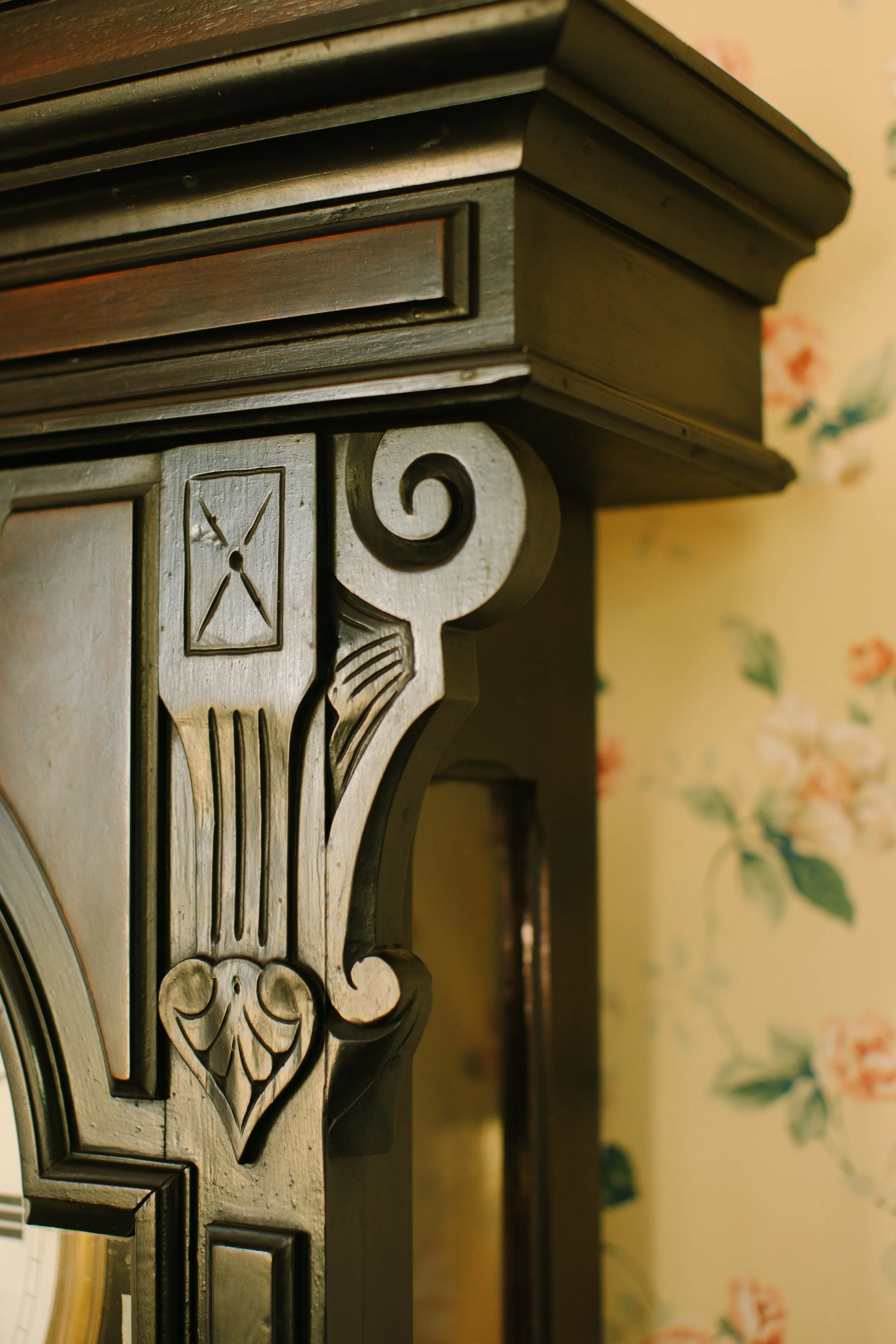 Close-up of an ornate wooden furniture corner with decorative carvings, against a floral-patterned wallpaper background.