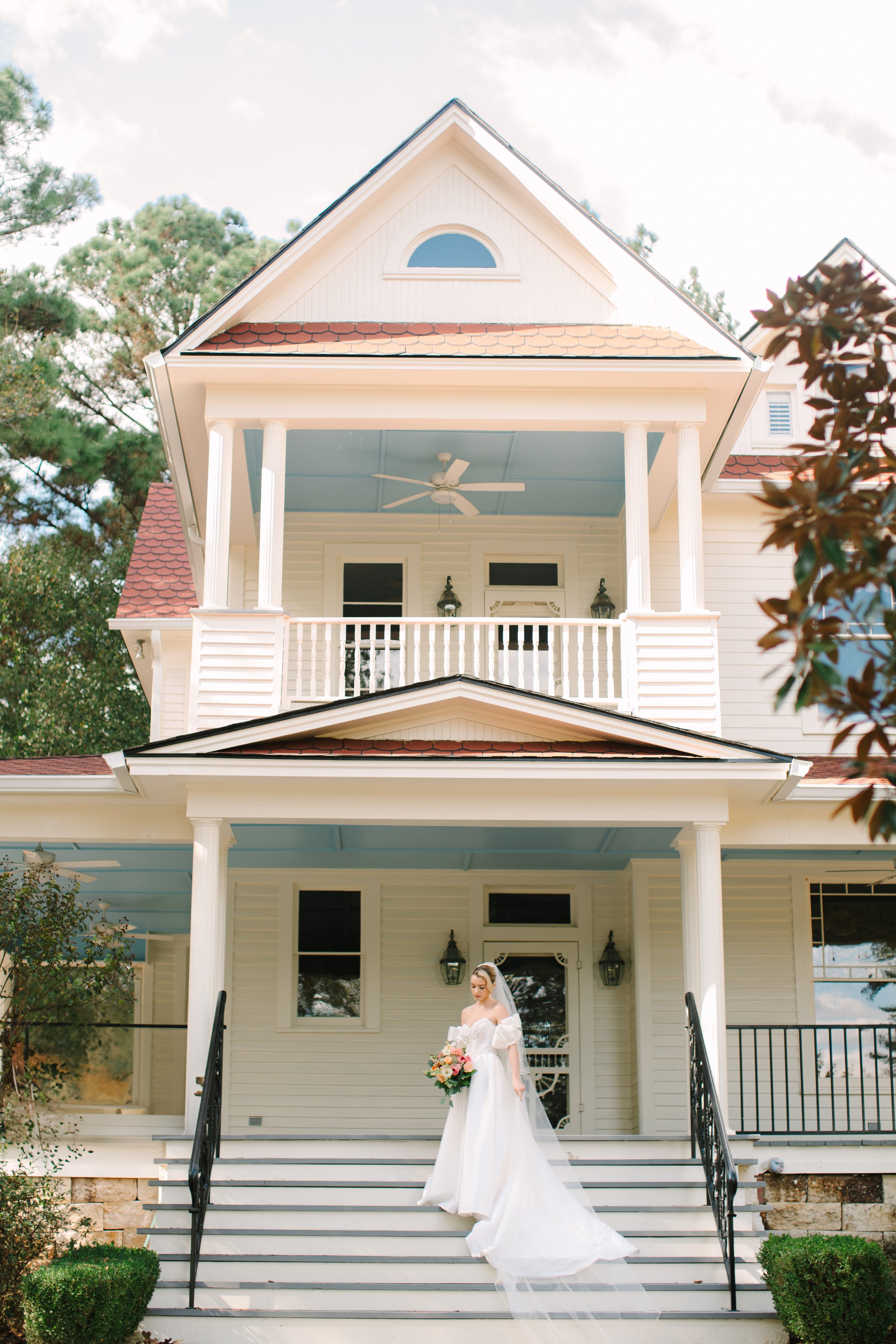 Bride in a white wedding gown with a train holding a bouquet of flowers on the steps of a large, white, three-story house with a porch and balcony.