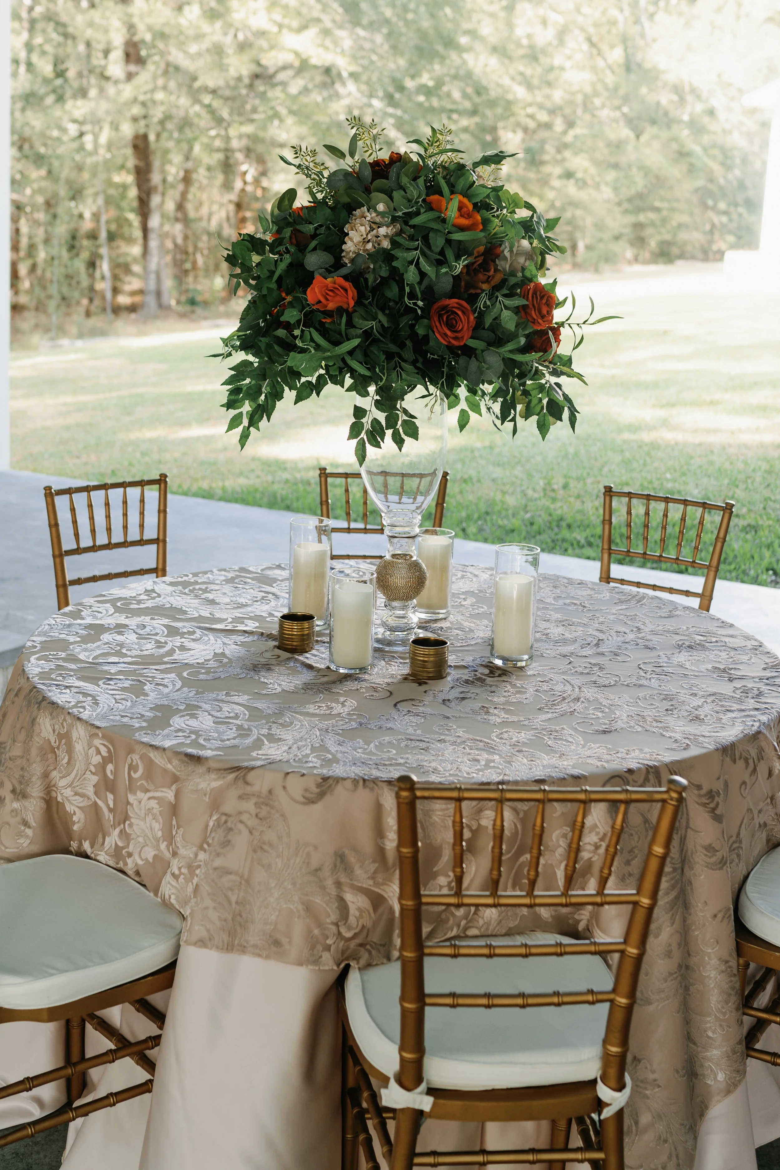 Round outdoor table with a floral centerpiece, candles, and decorative accents, set for an event with gold chairs and a green outdoor background.