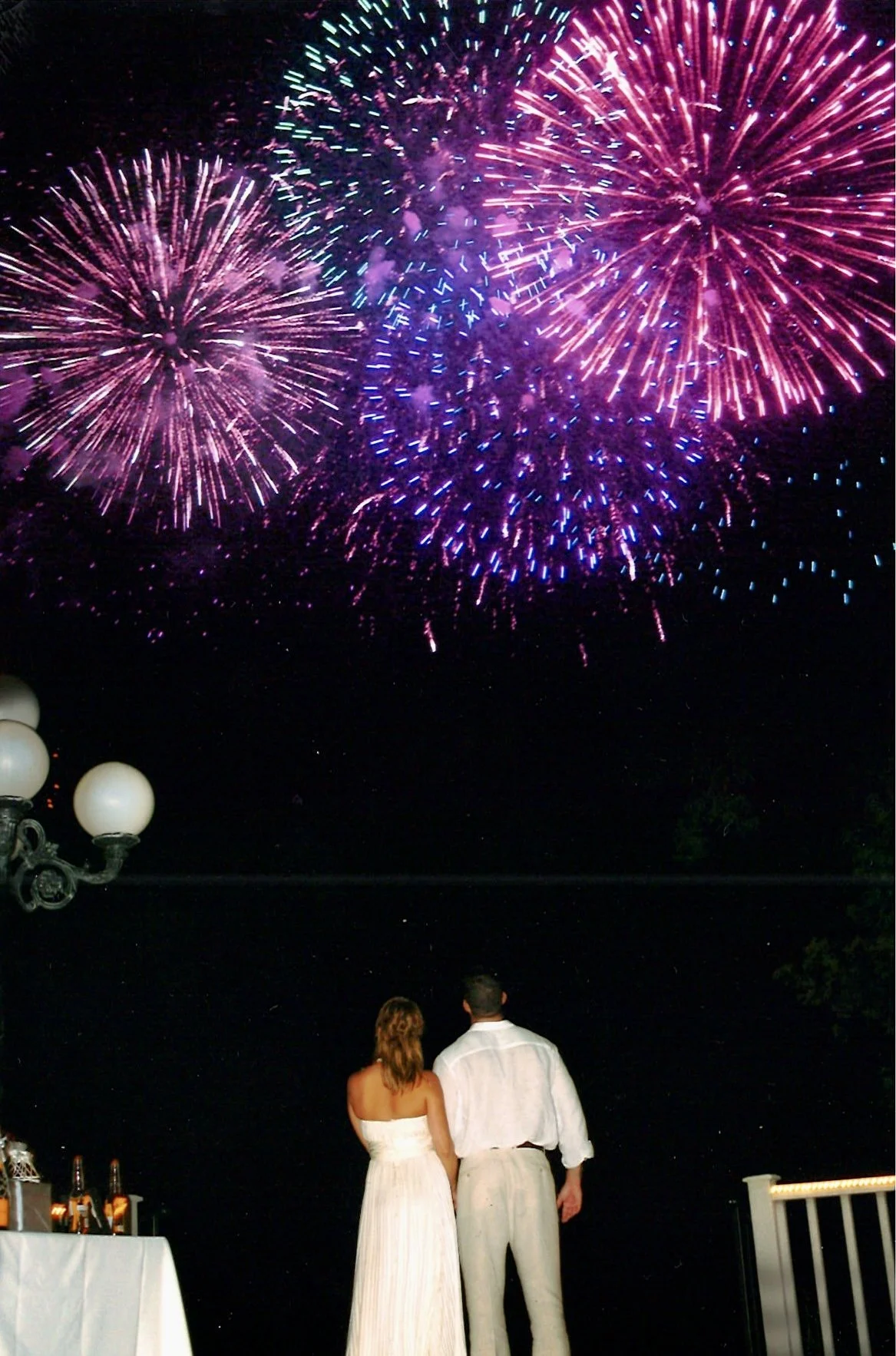 A couple dressed in formal attire watching colorful fireworks display in the night sky.
