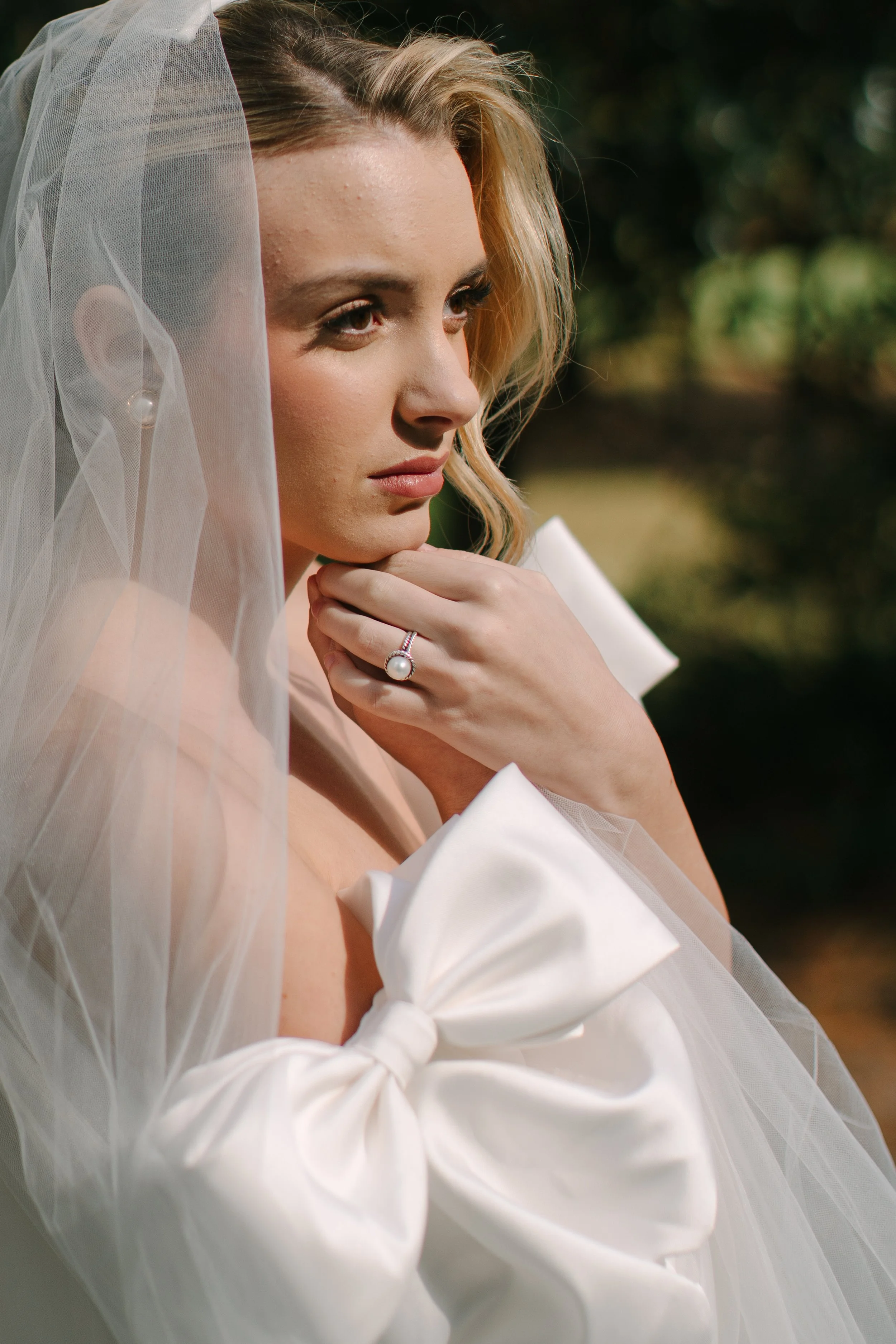 A bride with blonde hair wearing a bridal veil, pearl earrings, and a ring, posing outdoors with greenery in the background.