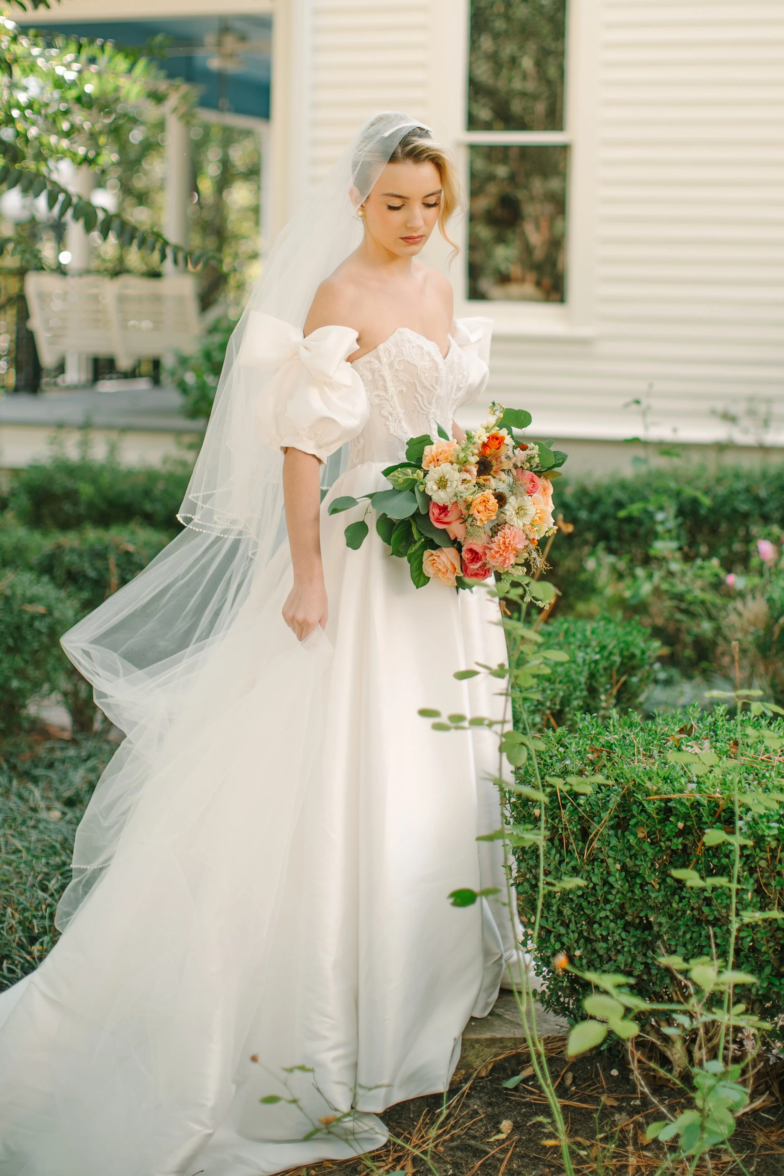 A bride in a white wedding gown holding a bouquet of colorful flowers stands outdoors near bushes with a white house in the background.