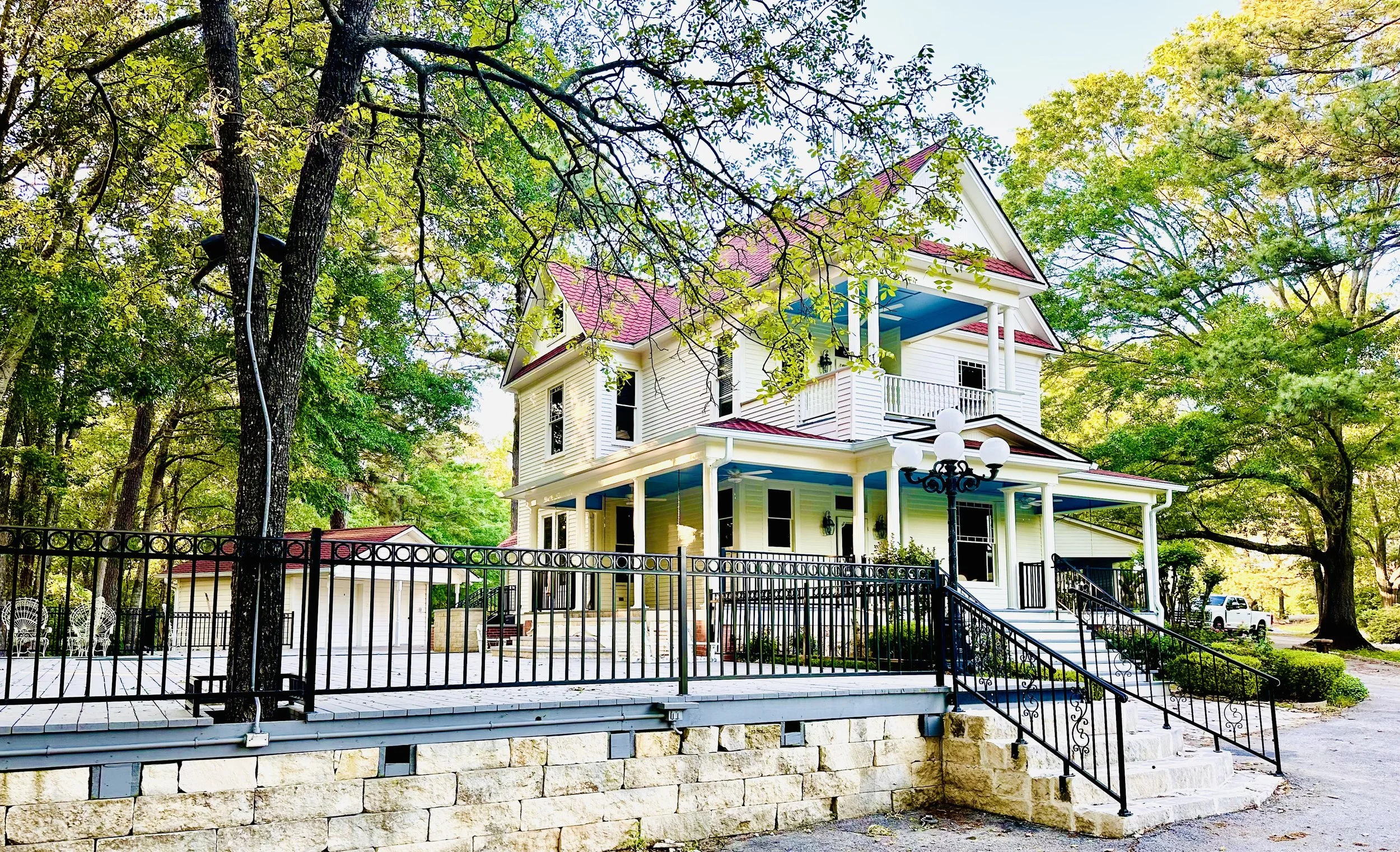 A large, white, three-story house with a red roof, surrounded by trees with green foliage, a black metal fence, and a set of stairs leading to the front porch.