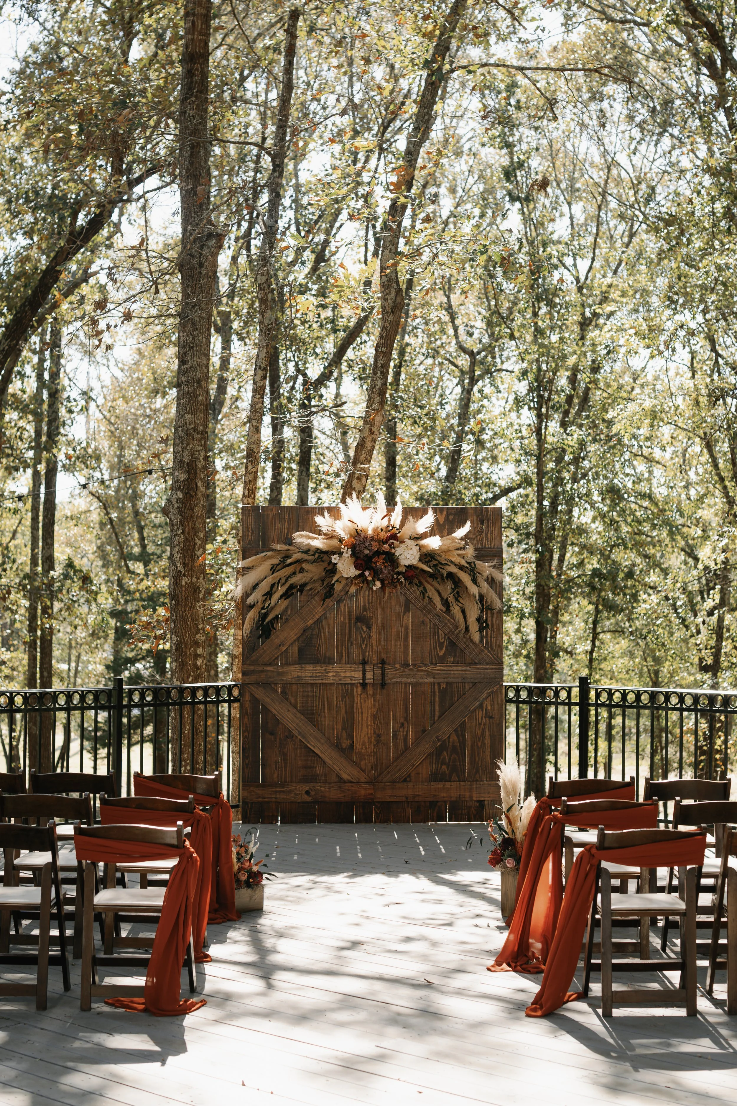 Outdoor wedding ceremony setup with wooden chairs adorned with orange sashes, facing a wooden arch decorated with dried flowers and pampas grass, surrounded by trees in a forest setting.